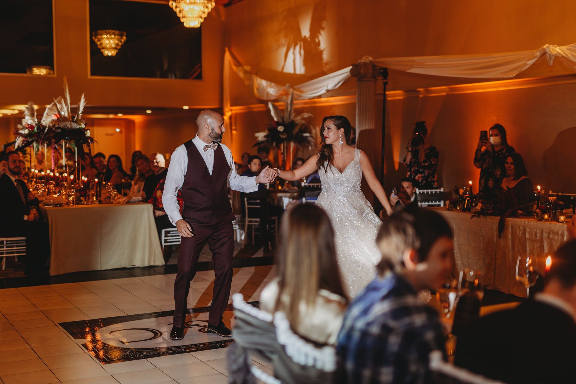 Couple dancing at a wedding reception; warm lighting, people watching, tables set.