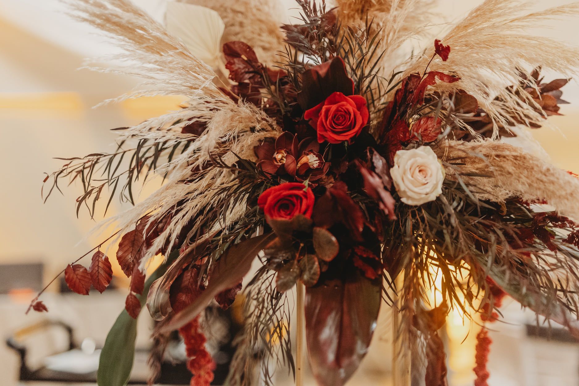 Floral arrangement: red and white roses, brown leaves, pampas grass, warm-toned, table setting.