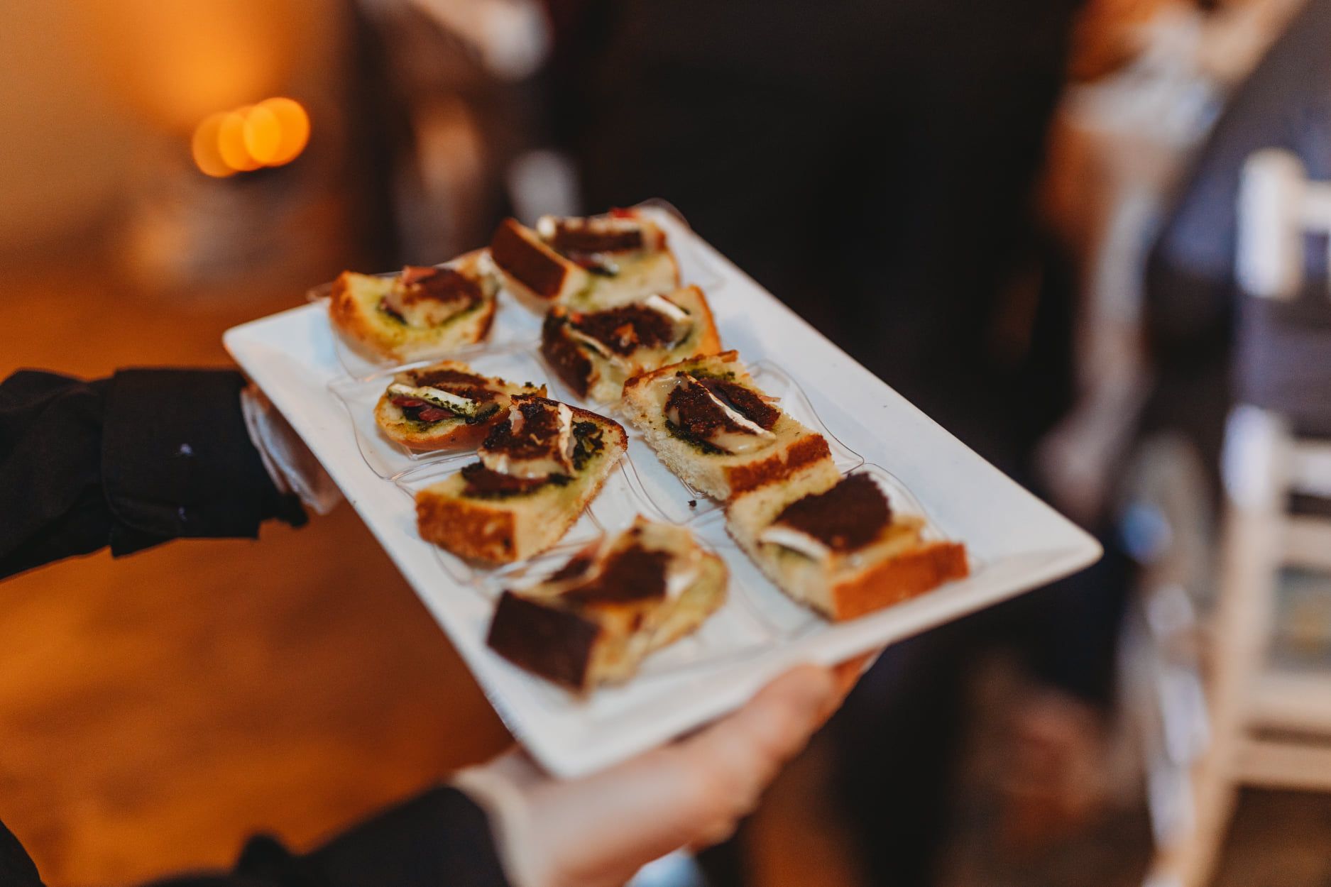 A server holding a rectangular plate of crostini appetizers, topped with dark spread and seasonings.