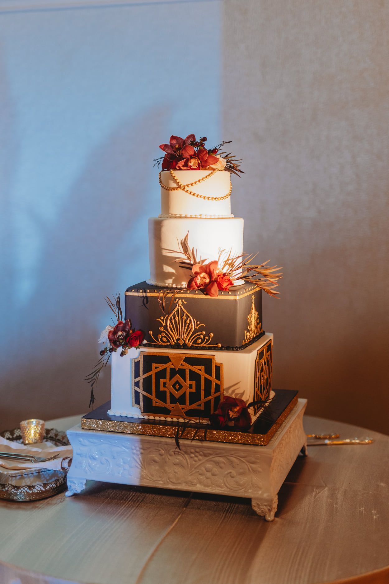 Four-tiered Art Deco-style wedding cake with gold accents and red flowers on a white square stand.