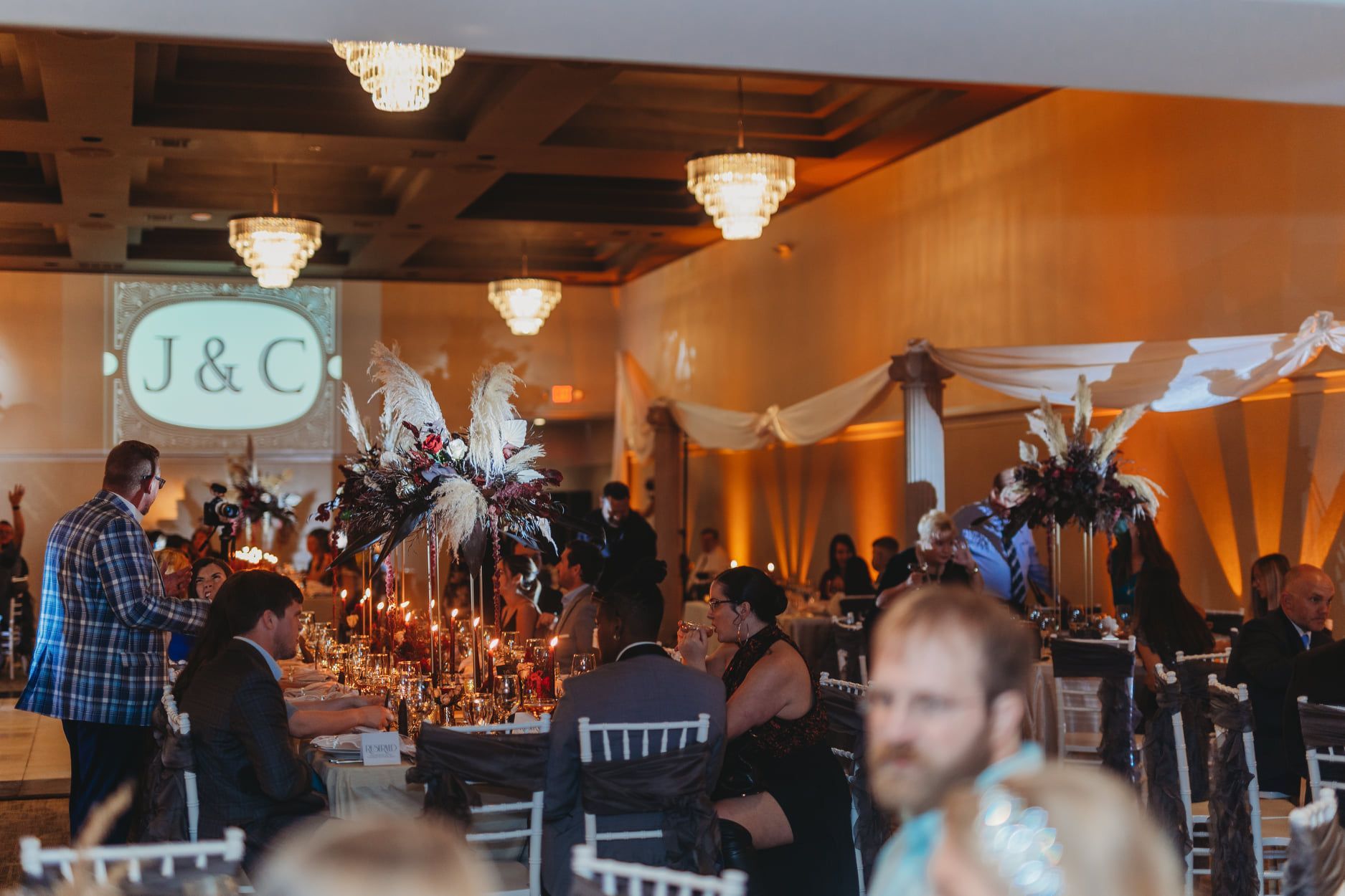 Wedding reception with guests seated at tables; floral centerpieces, monogram projected on wall.