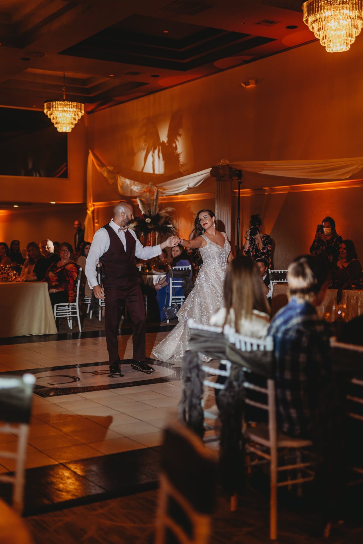 Bride and groom dancing at wedding reception. Groom points, bride smiles in sparkly gown. Guests watch, band on stage.
