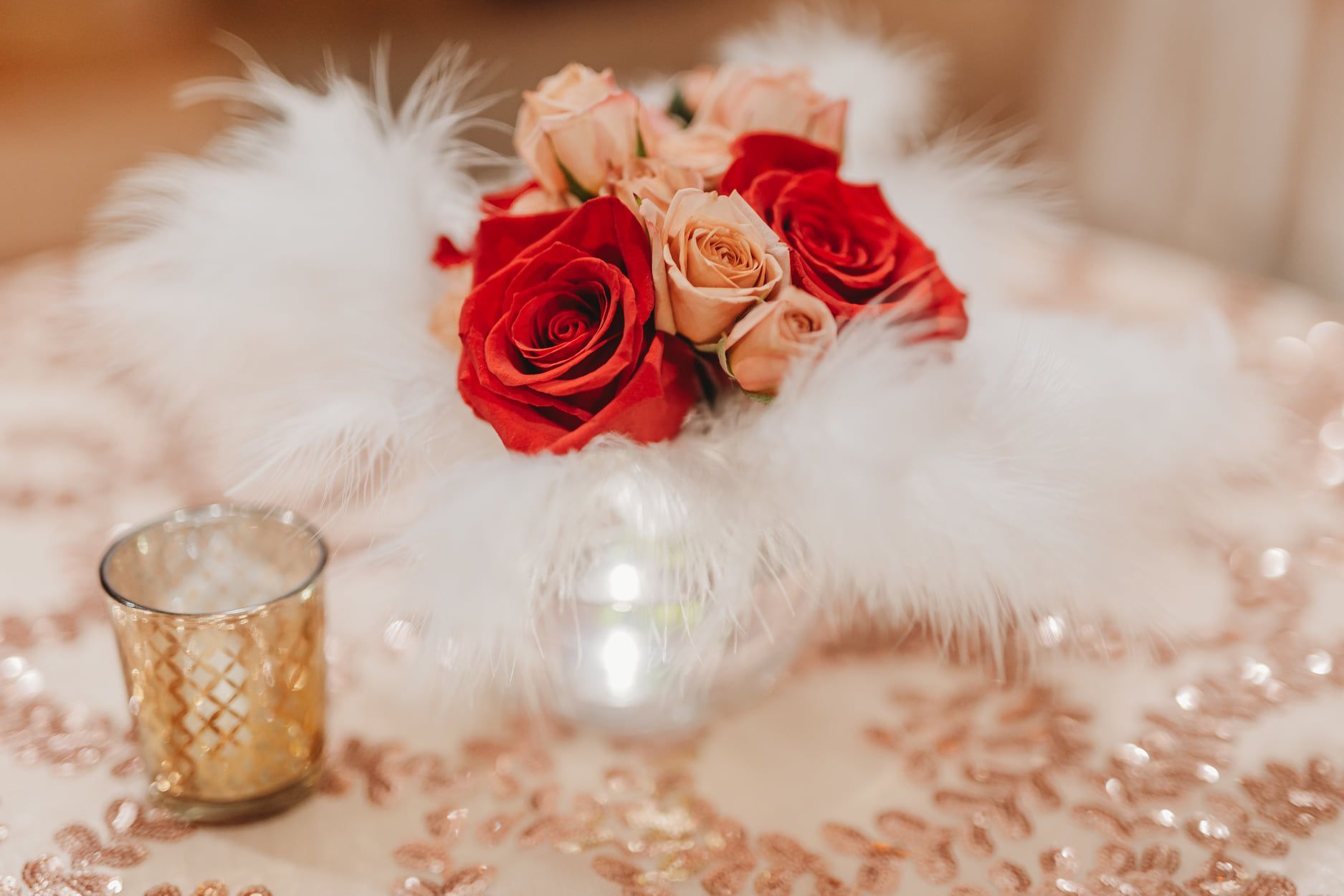 Red and peach roses atop white feathers, on a rose-gold sequined tablecloth, with a gold votive.