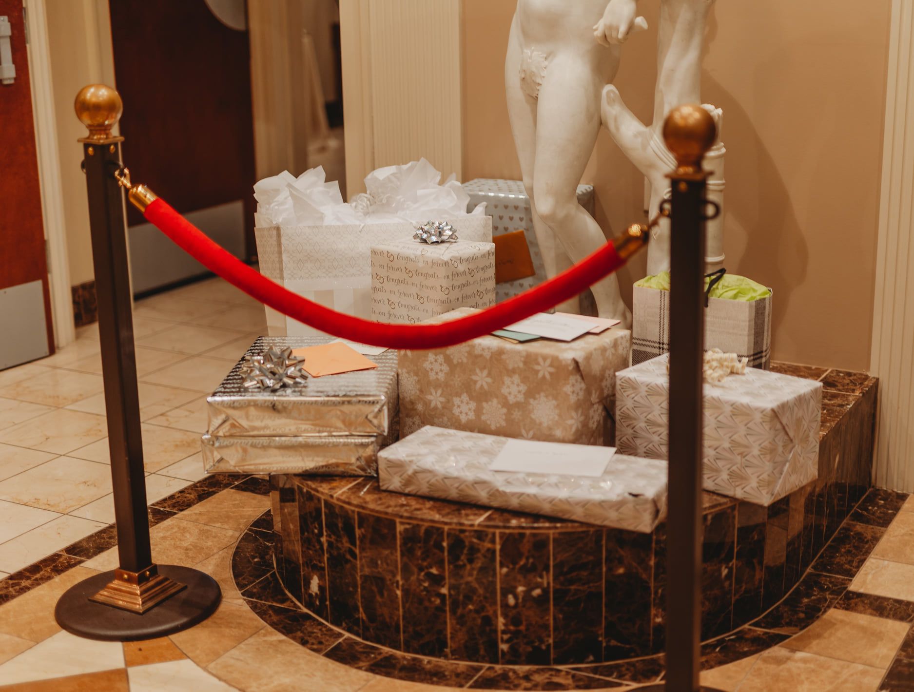 Gifts wrapped in various papers on a marble pedestal, cordoned off with a red velvet rope and stanchions.