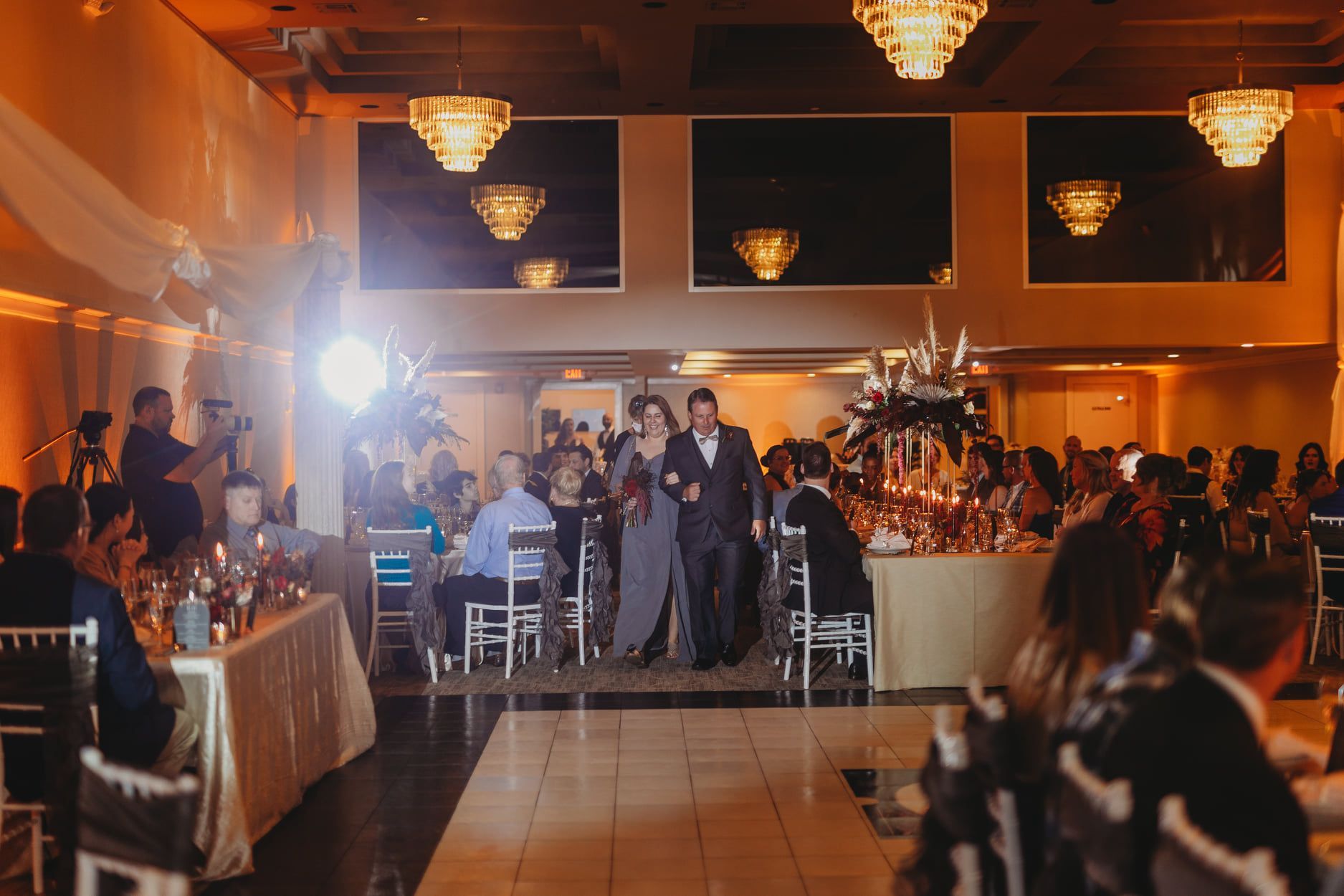 Wedding reception with guests and a couple walking down the aisle. Warm lighting, chandeliers, and floral centerpieces.
