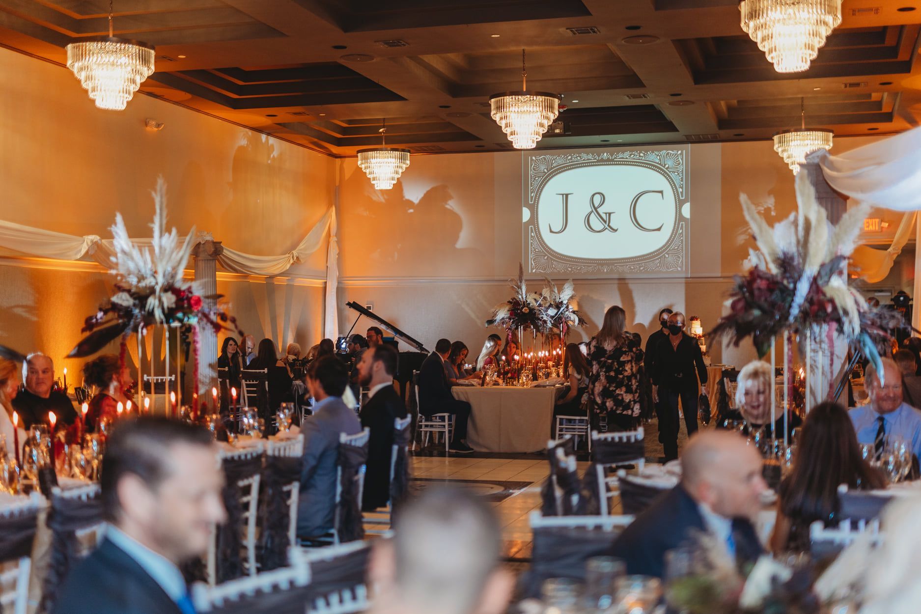 Wedding reception with guests seated at tables; floral centerpieces, initials projected on screen.