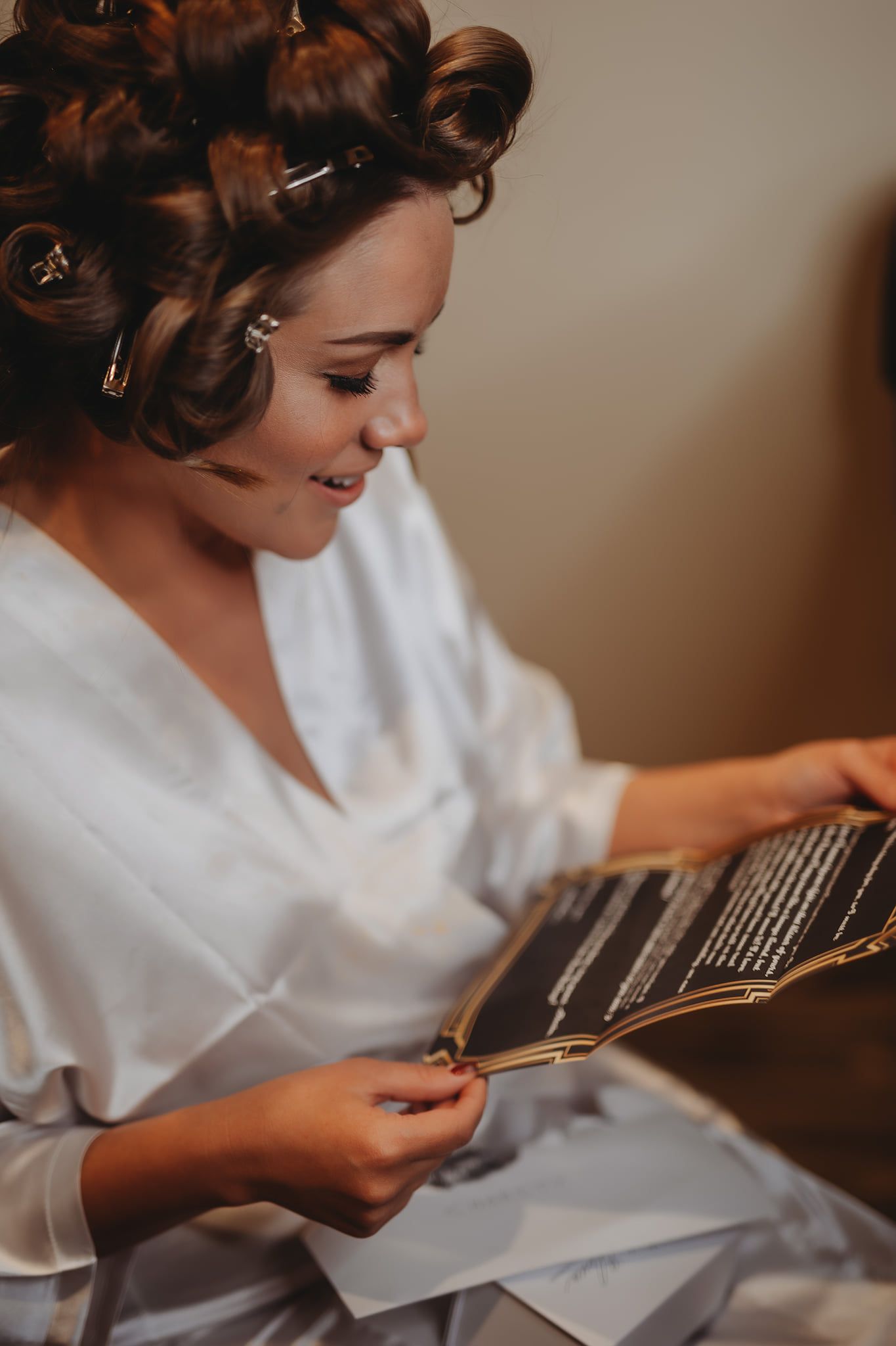 Woman in hair rollers, robe, smiling, looking at a folded black and gold fan, indoors.