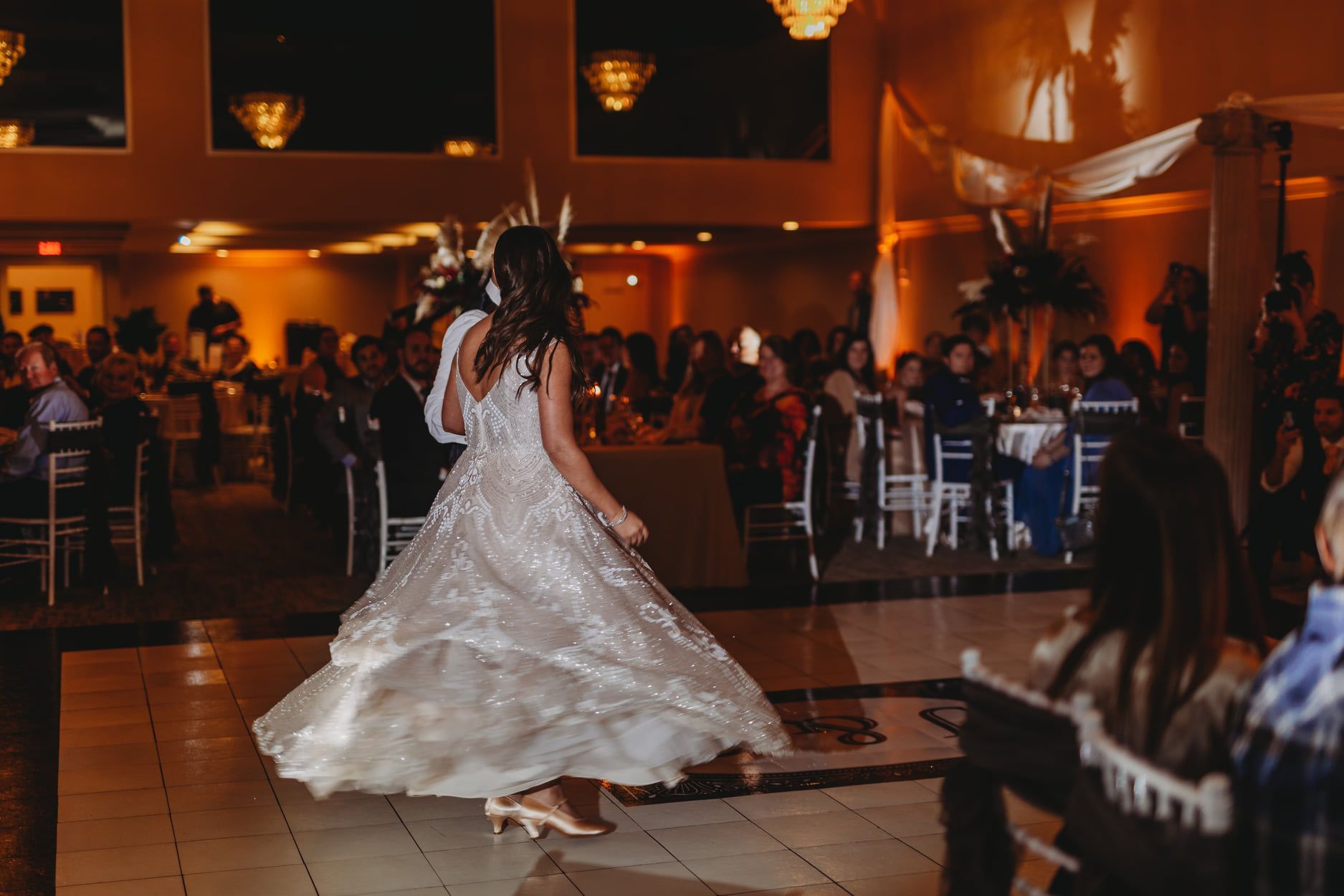 Bride dancing at a wedding reception, in a sparkling gown, surrounded by guests at tables.