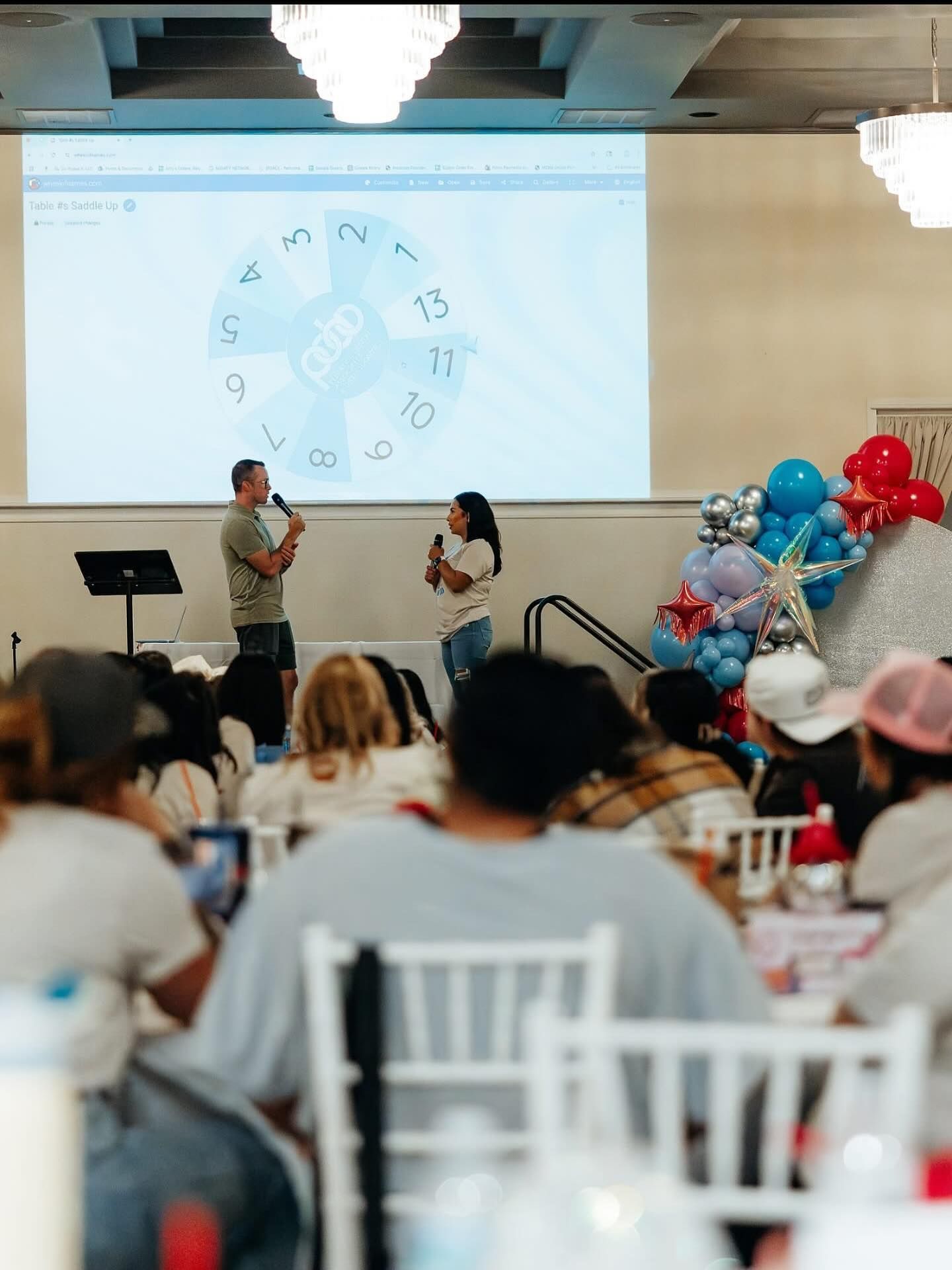 Two people on stage with microphones, in front of a screen with a prize wheel; audience in chairs.