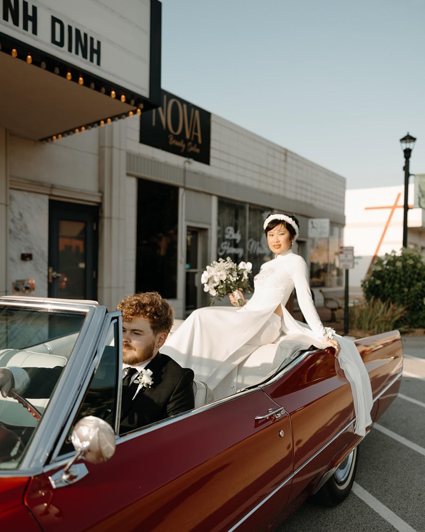 Bride in white dress poses on a red convertible with groom driving, outside a building with 