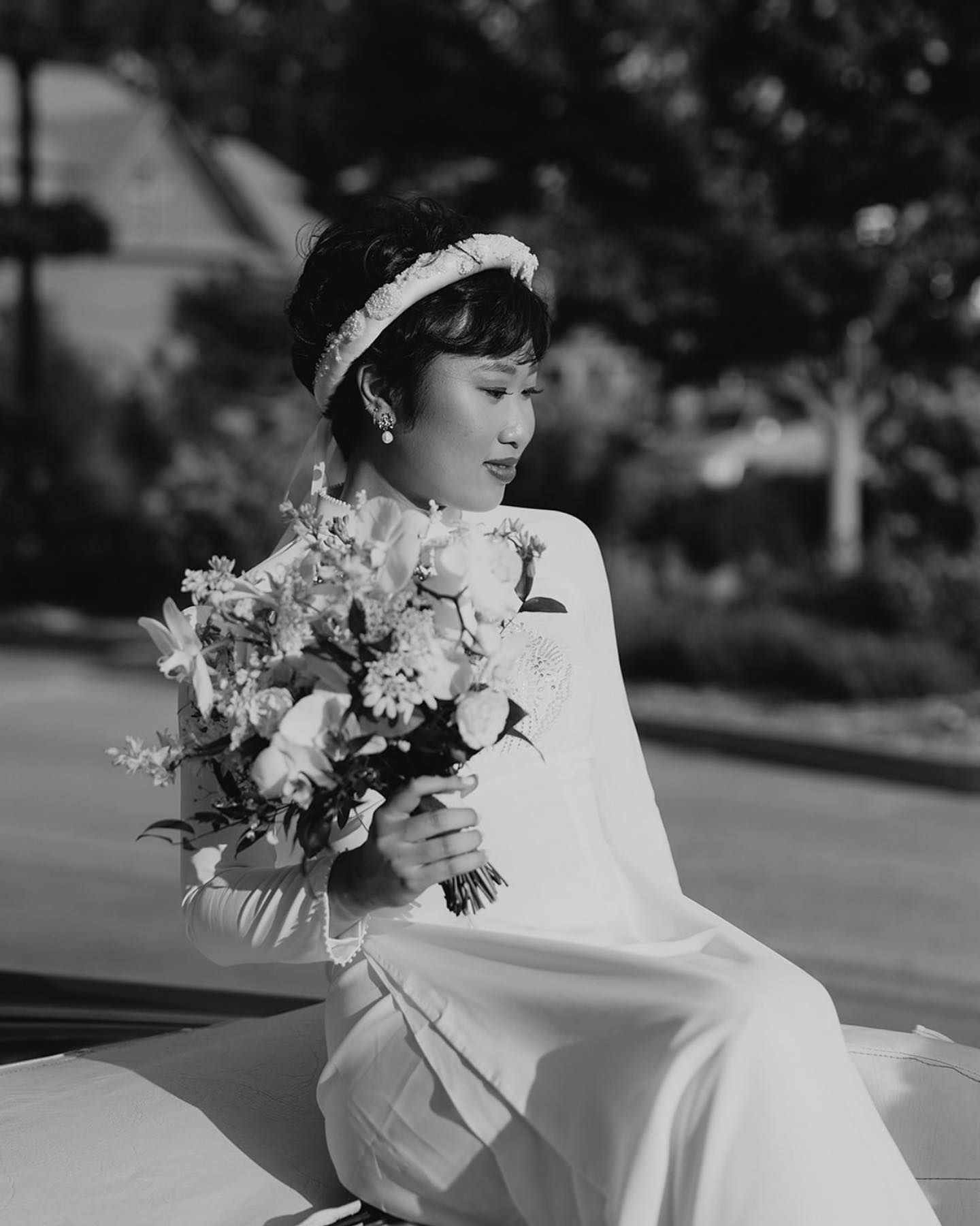 Woman in white dress and floral headpiece holding flowers, sitting outside.