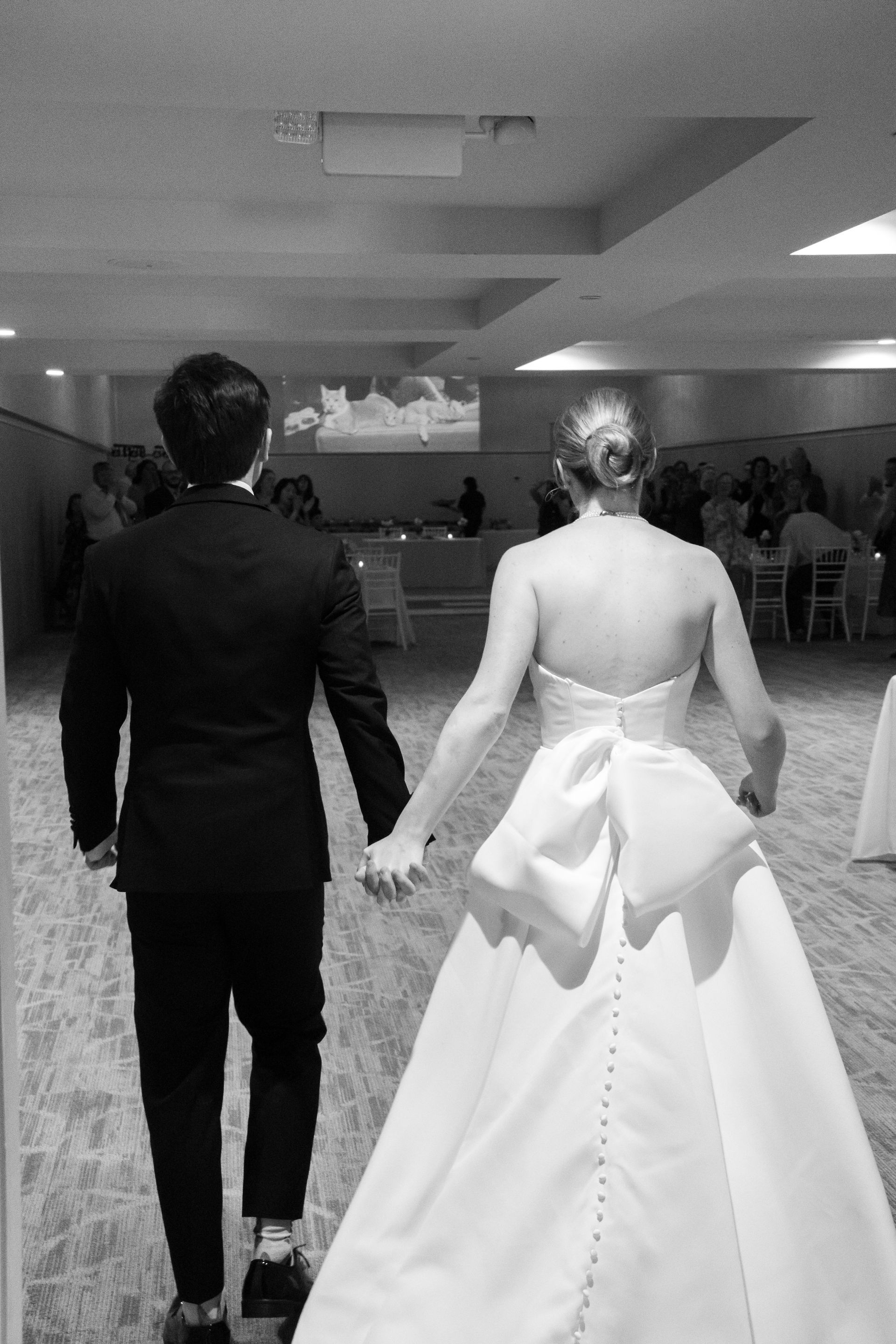 Newlyweds hold hands walking into reception. Bride in white gown, groom in suit.