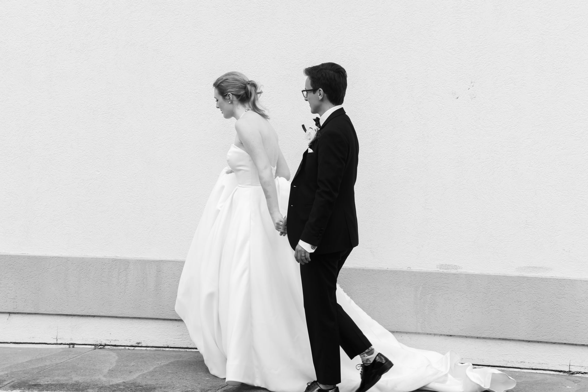 Bride and groom walk hand-in-hand. She wears a white gown, and he wears a black suit. Against a white wall.