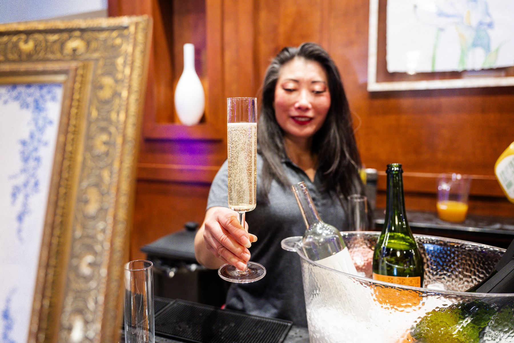 Woman holding a glass of champagne, offering it forward. She is in a room with a painting and bar setup.