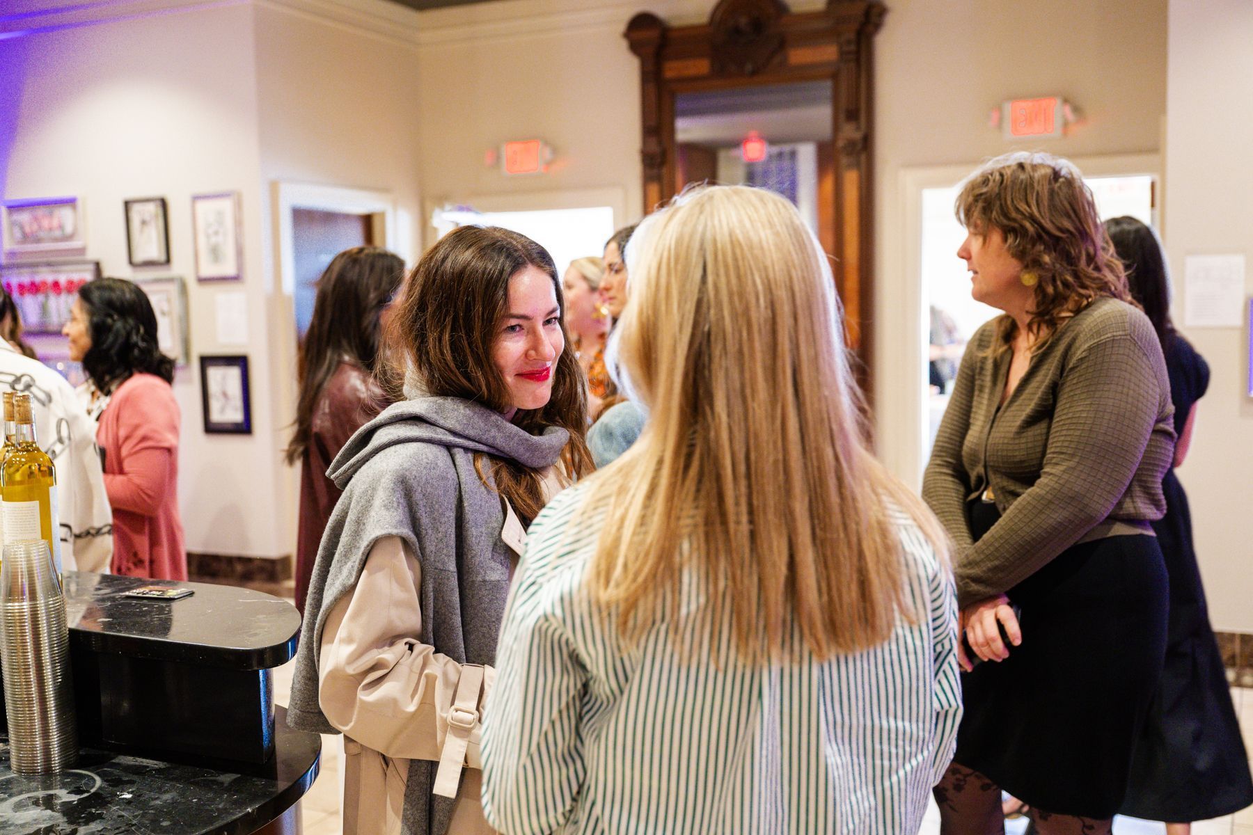 People socializing in a well-lit interior. Two women converse, others look on. Artwork hangs on walls.