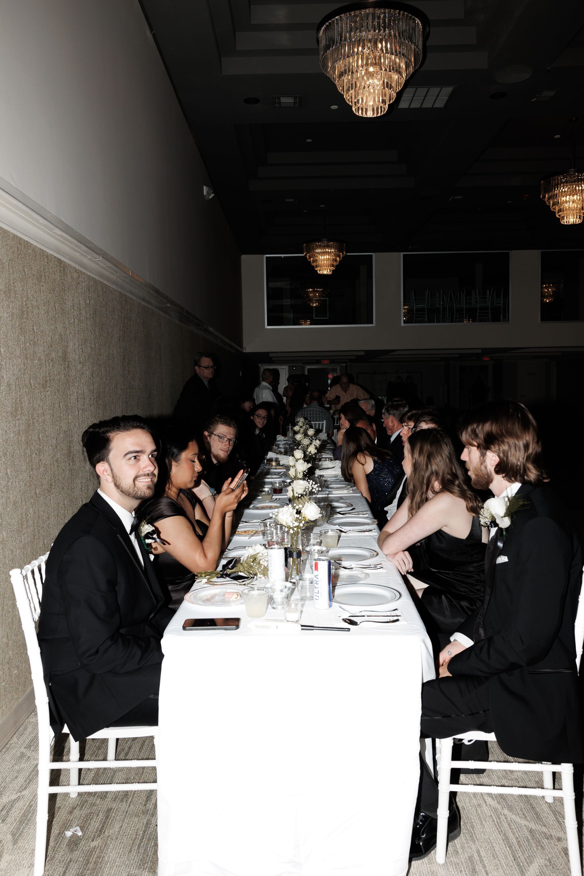 People seated at a long white table in a formal dining room, illuminated by chandeliers, engaged in conversation.