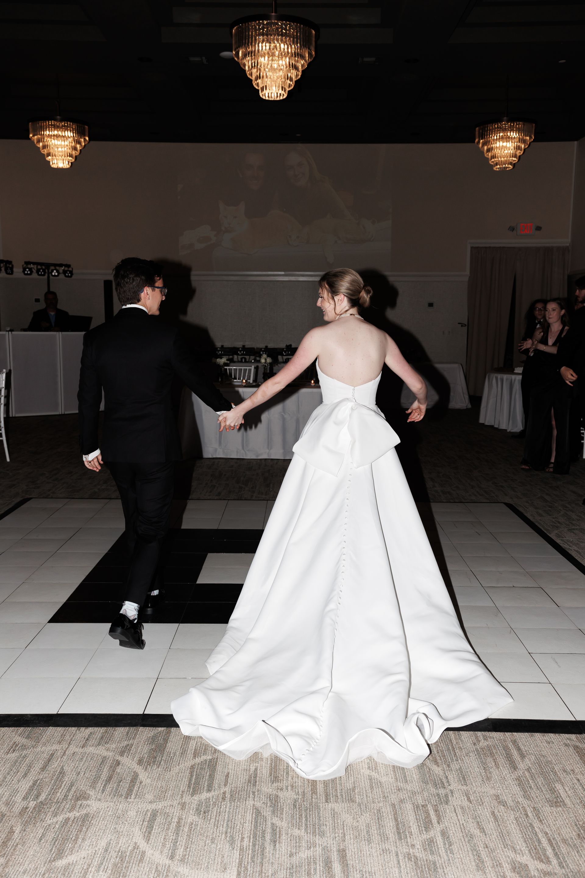 Bride and groom dance on a checkered dance floor at a reception. She wears a white gown, he wears a black tuxedo.