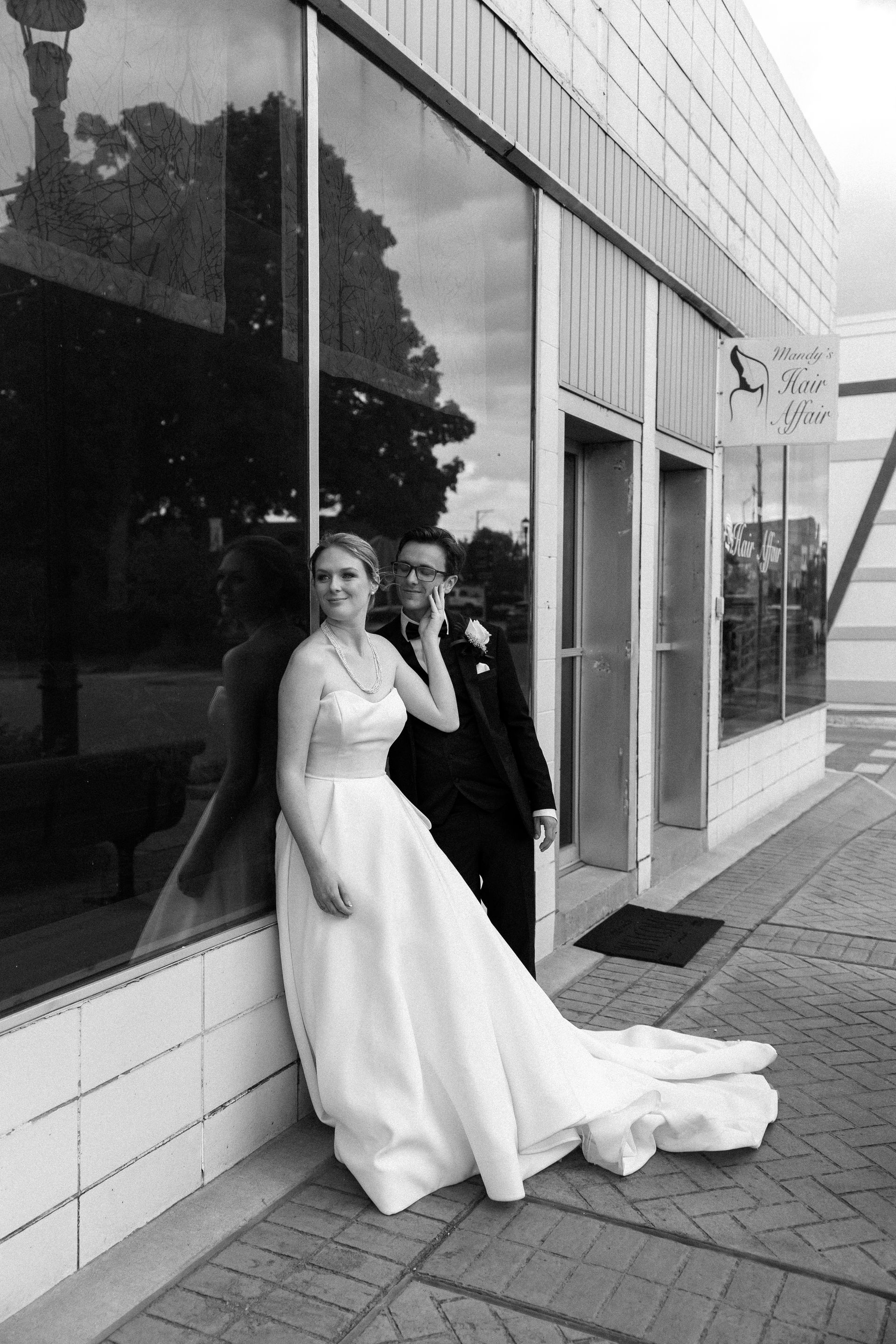 Bride and groom pose outside a building with large windows. The bride wears a gown, and the groom is in a suit.