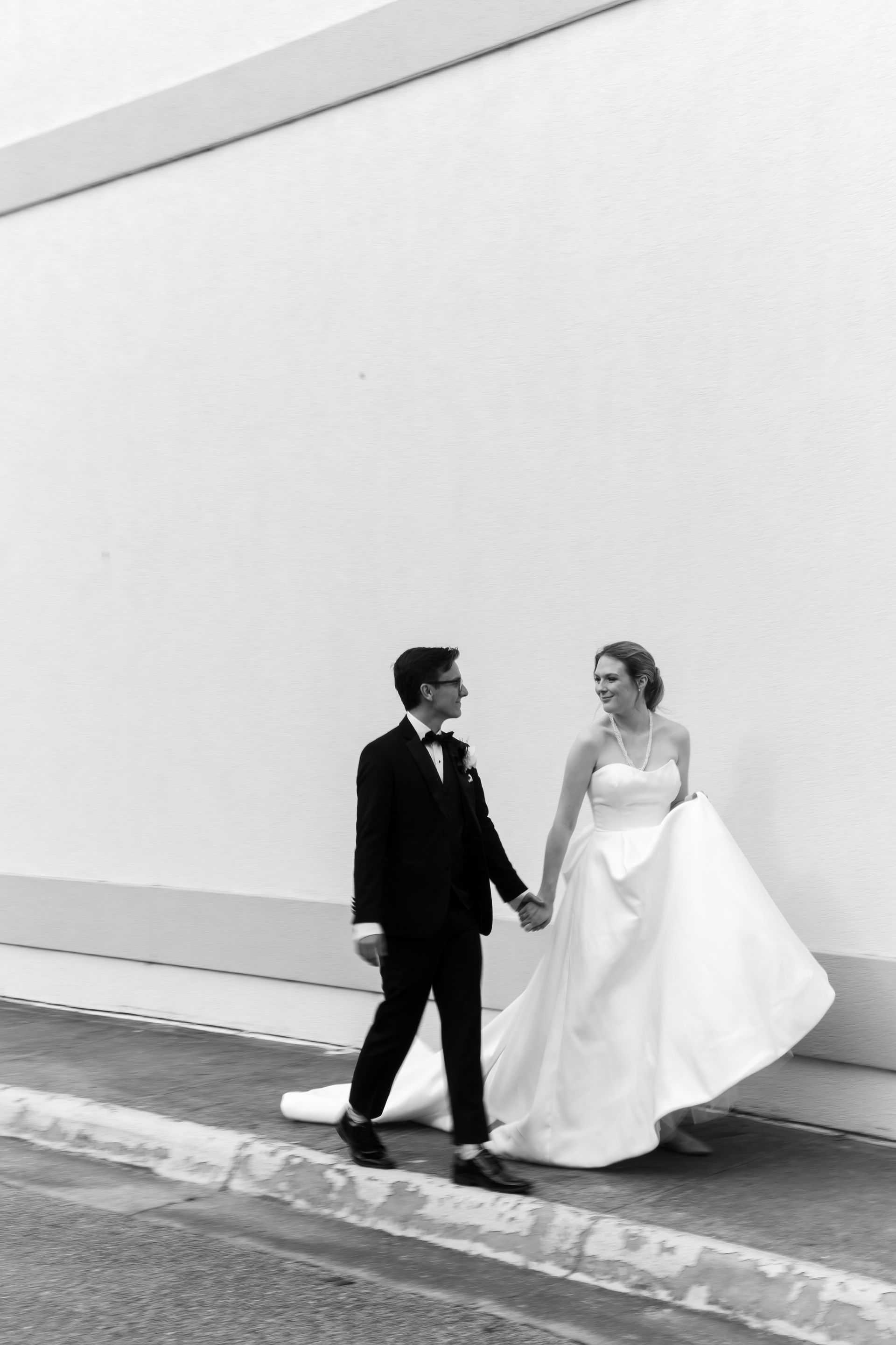 Bride and groom holding hands, walking on a sidewalk, smiling. Groom in tuxedo, bride in strapless gown, in front of a white wall.