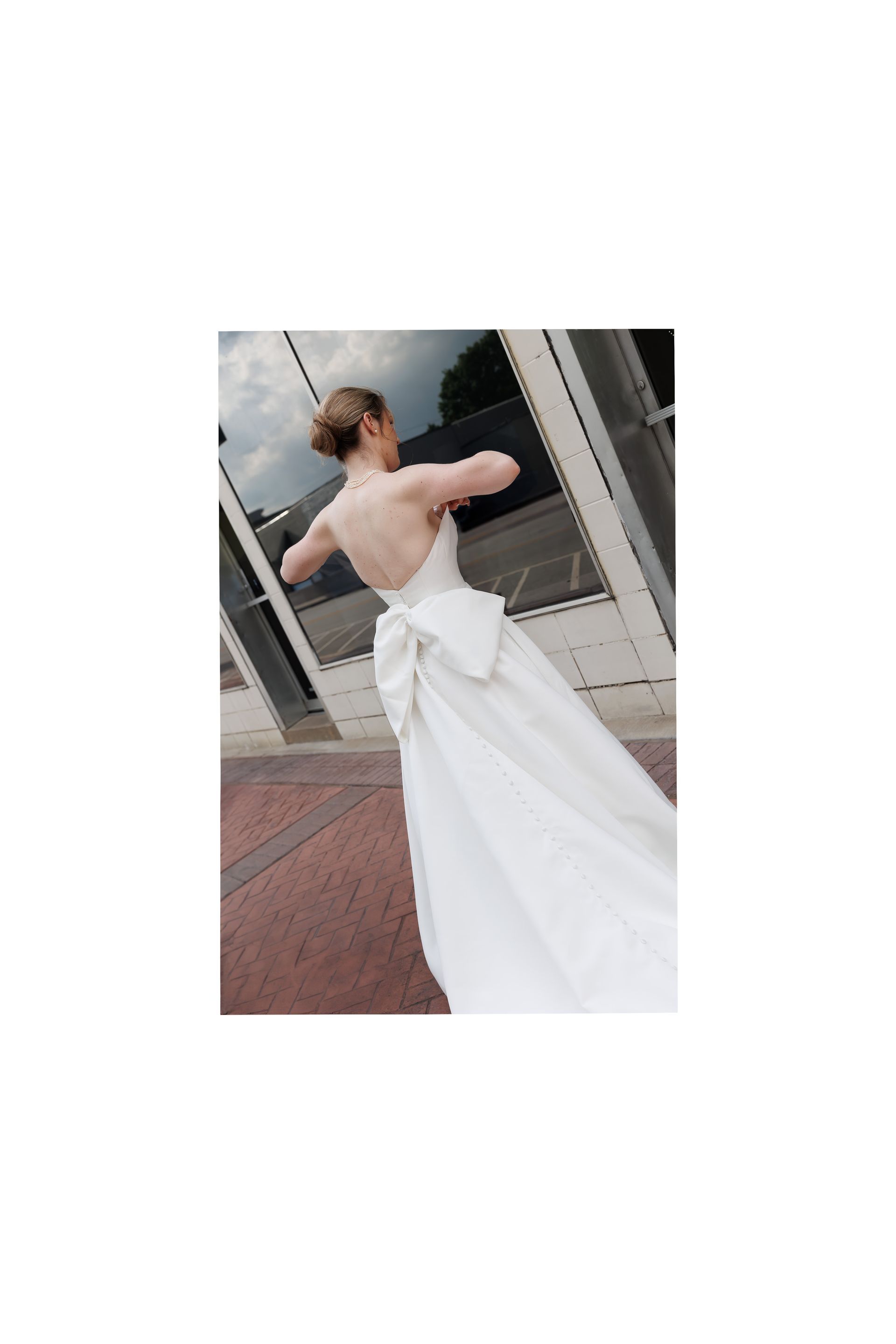 Woman in white wedding dress with bow, standing in front of glass windows.