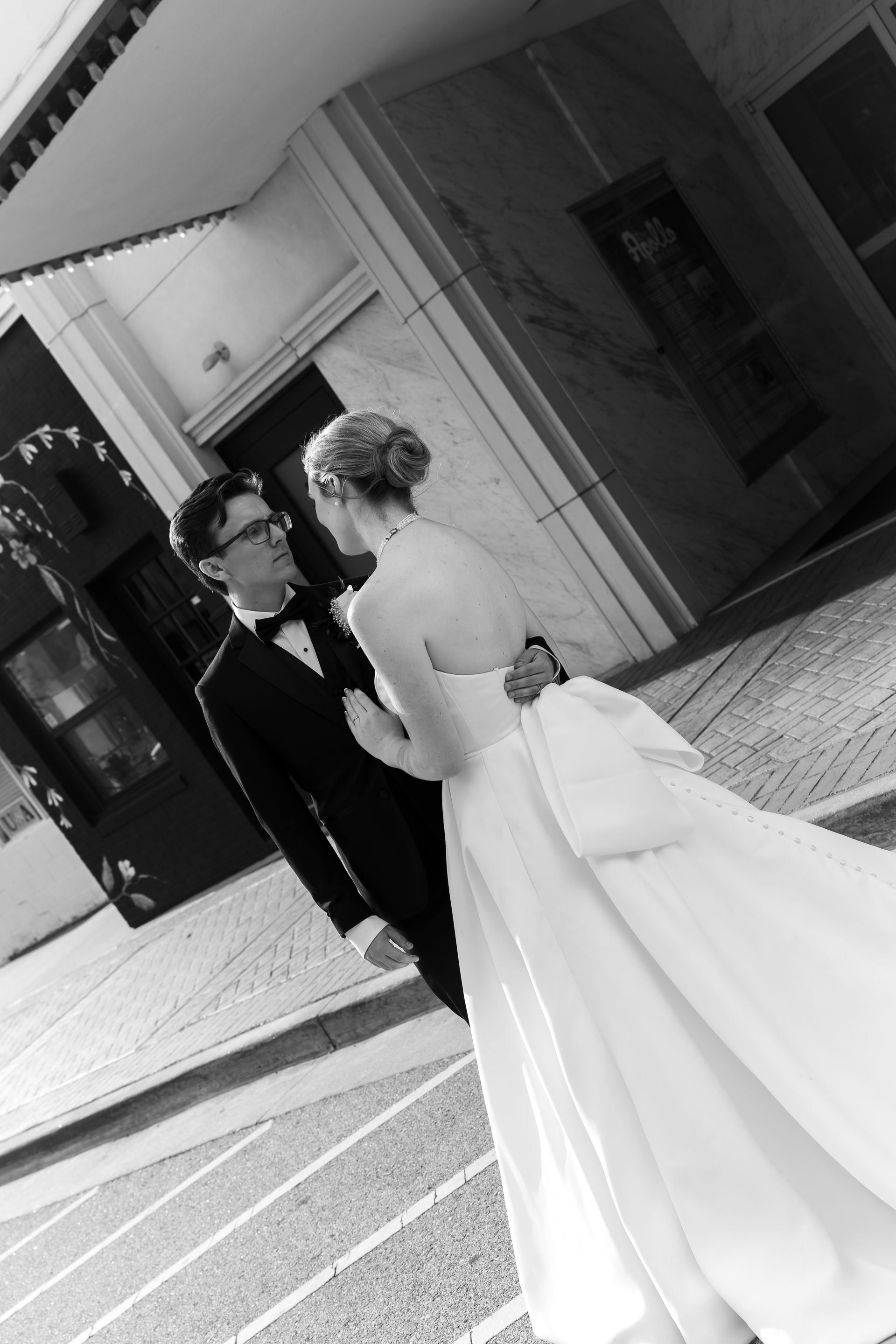 Bride and groom embrace in front of a building; black and white photograph.