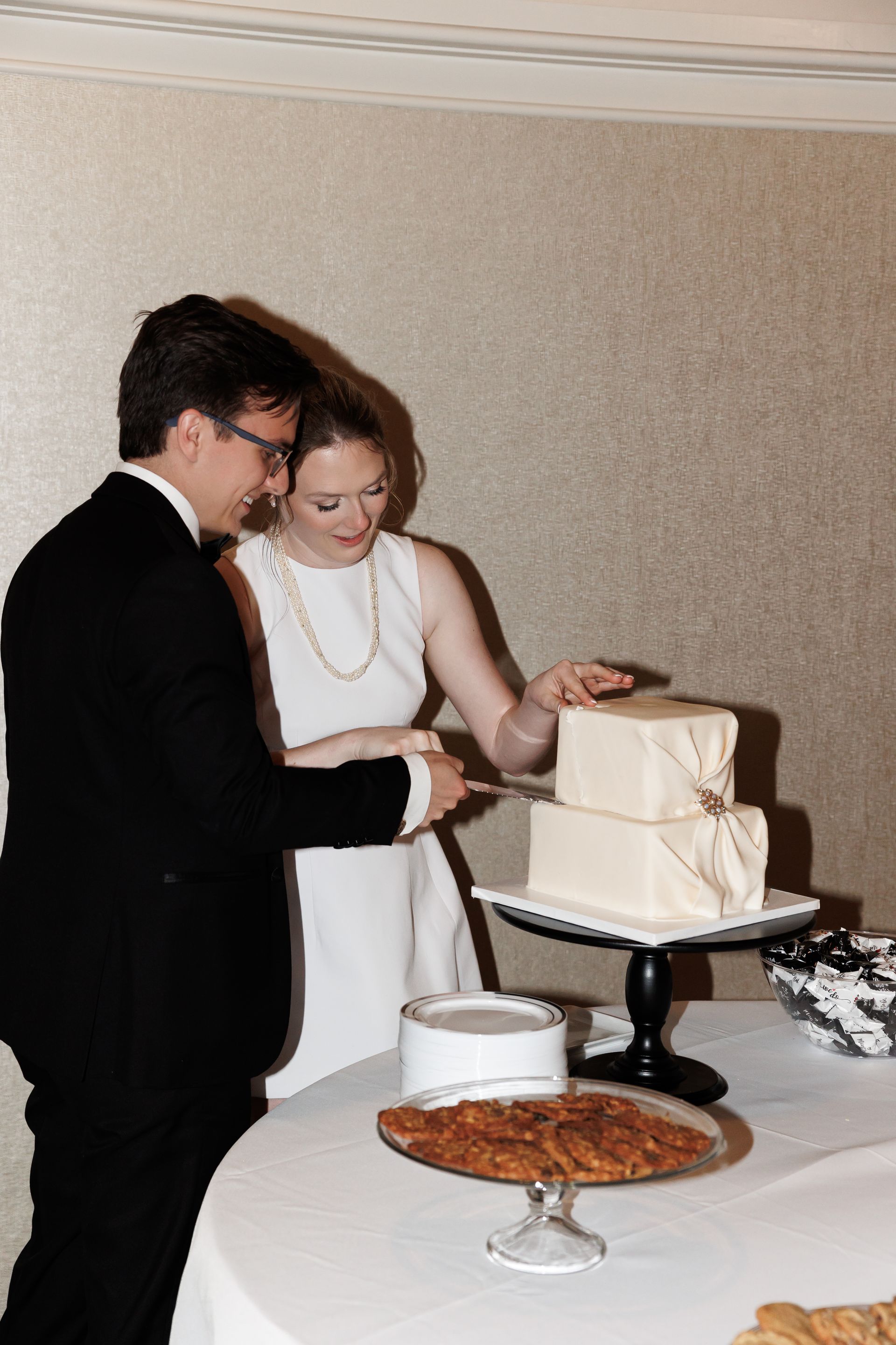 Couple cutting a two-tiered wedding cake. Man in black suit, woman in white dress. White cake with ribbon, on a black stand.