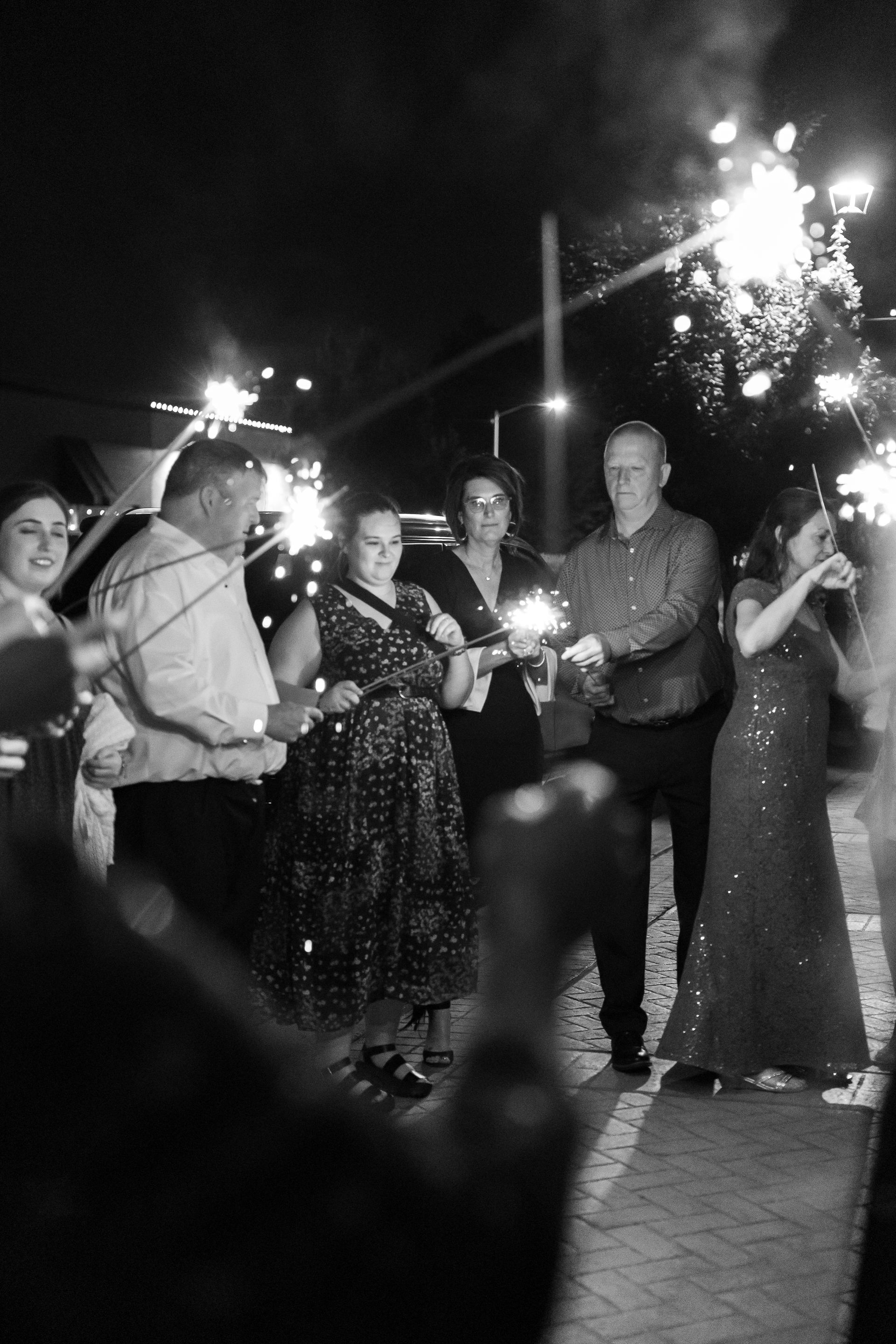 People holding sparklers outdoors at night, celebrating; fireworks in background.