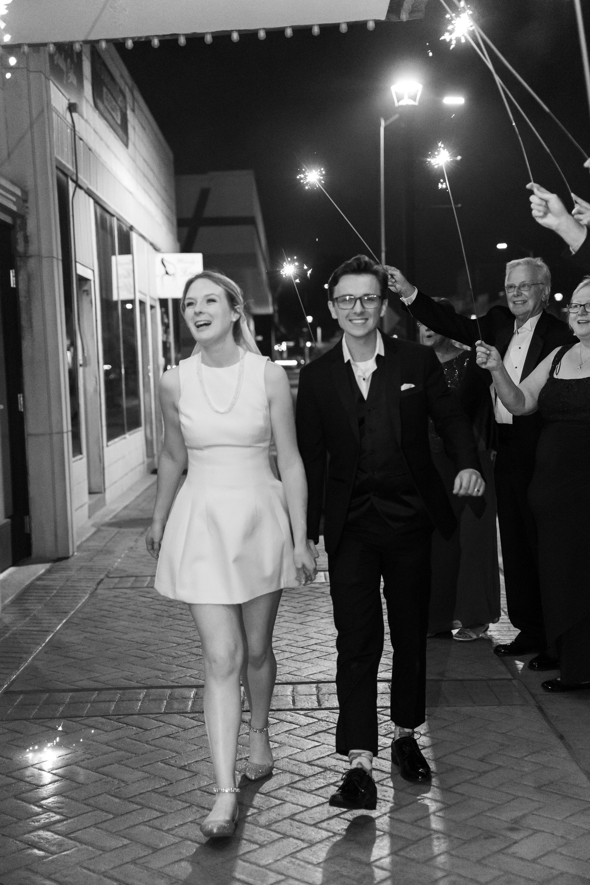 Couple walking, smiling, holding hands, illuminated by sparklers. Night exterior, building background.