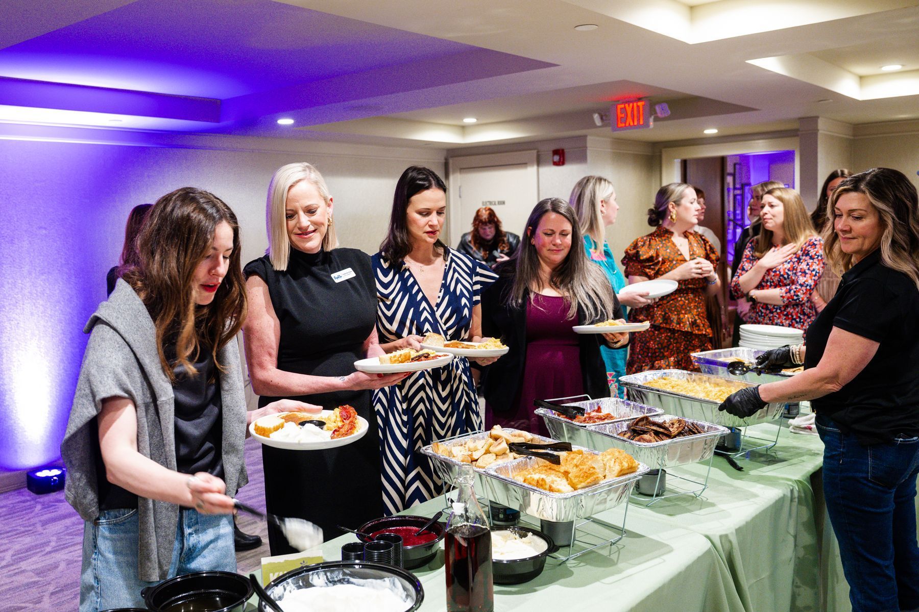 People at a buffet, serving themselves food. A person in black gloves assists.