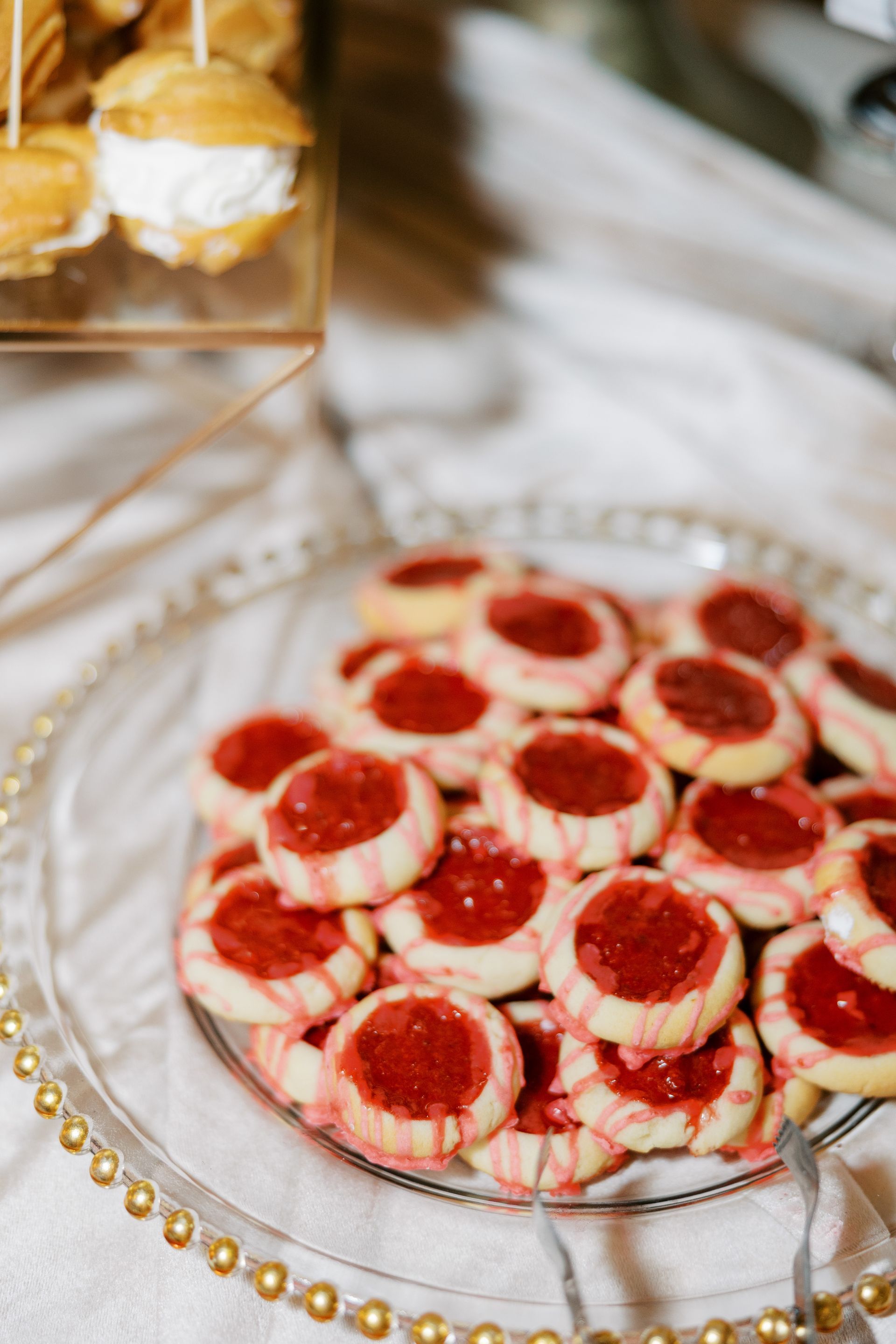 Cookies with red jam filling on a glass plate, cream puffs on top, white cloth background.