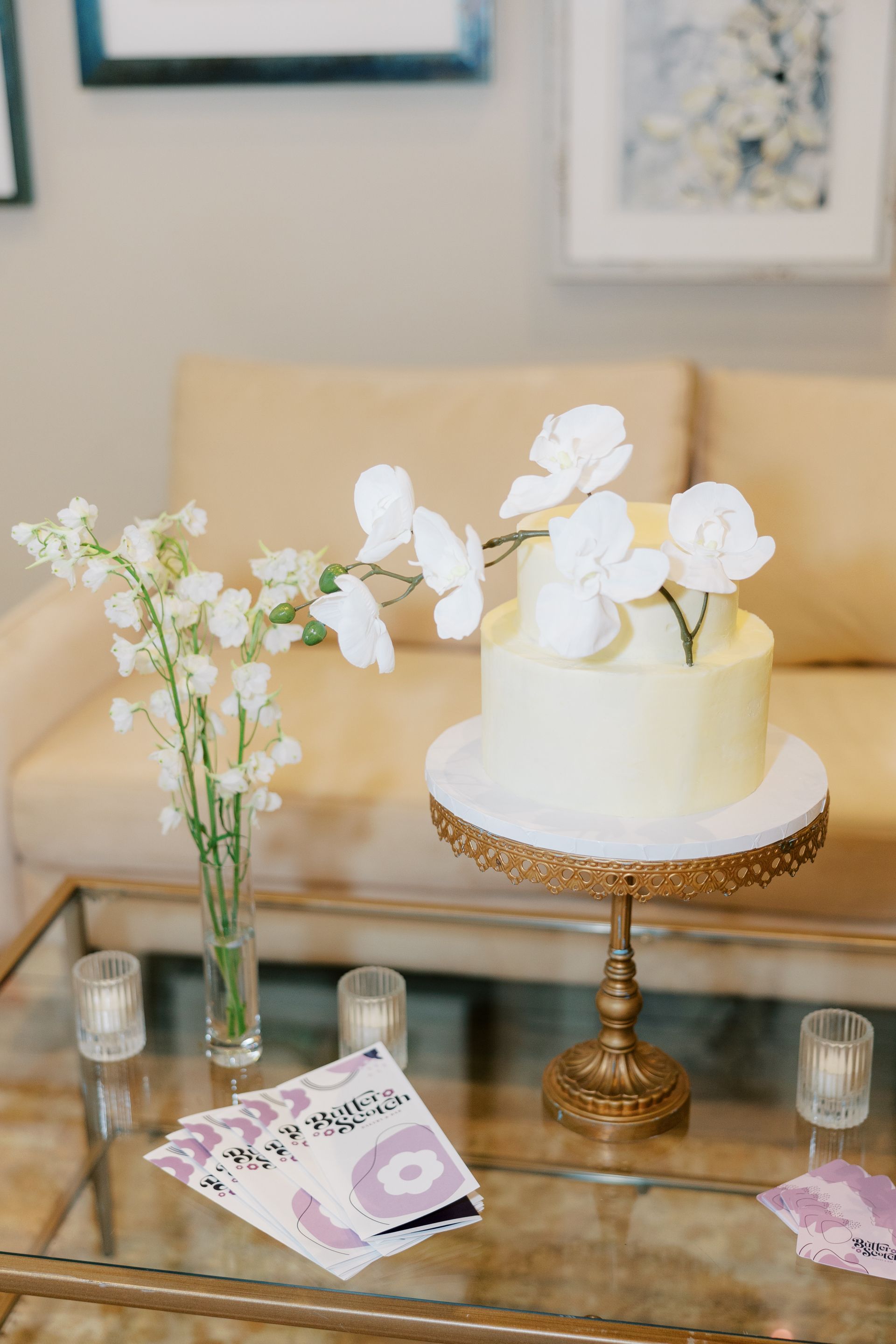 Two-tiered yellow cake with white flower decorations on a gold cake stand, next to flowers and cards on a glass table.