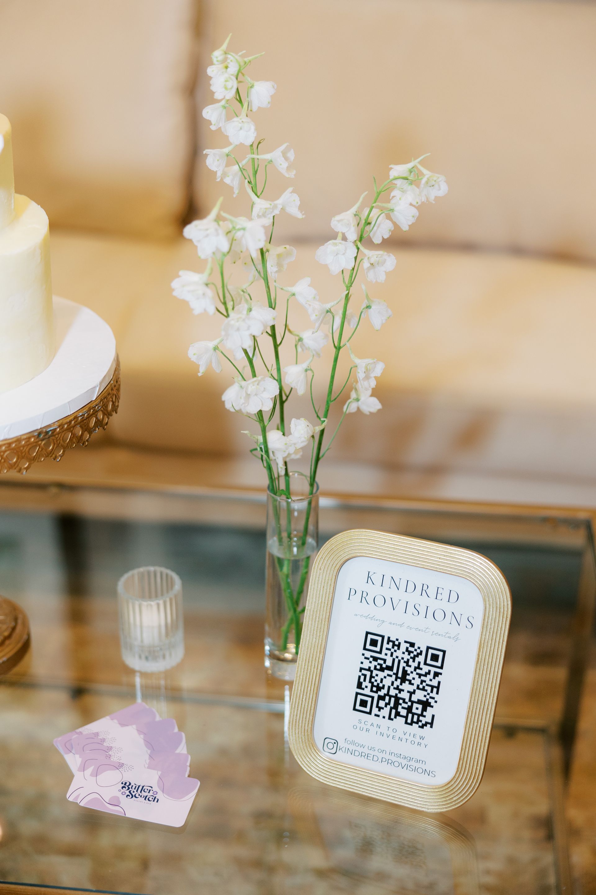 Clear glass vase with white flowers, gold framed QR code sign on a glass table.