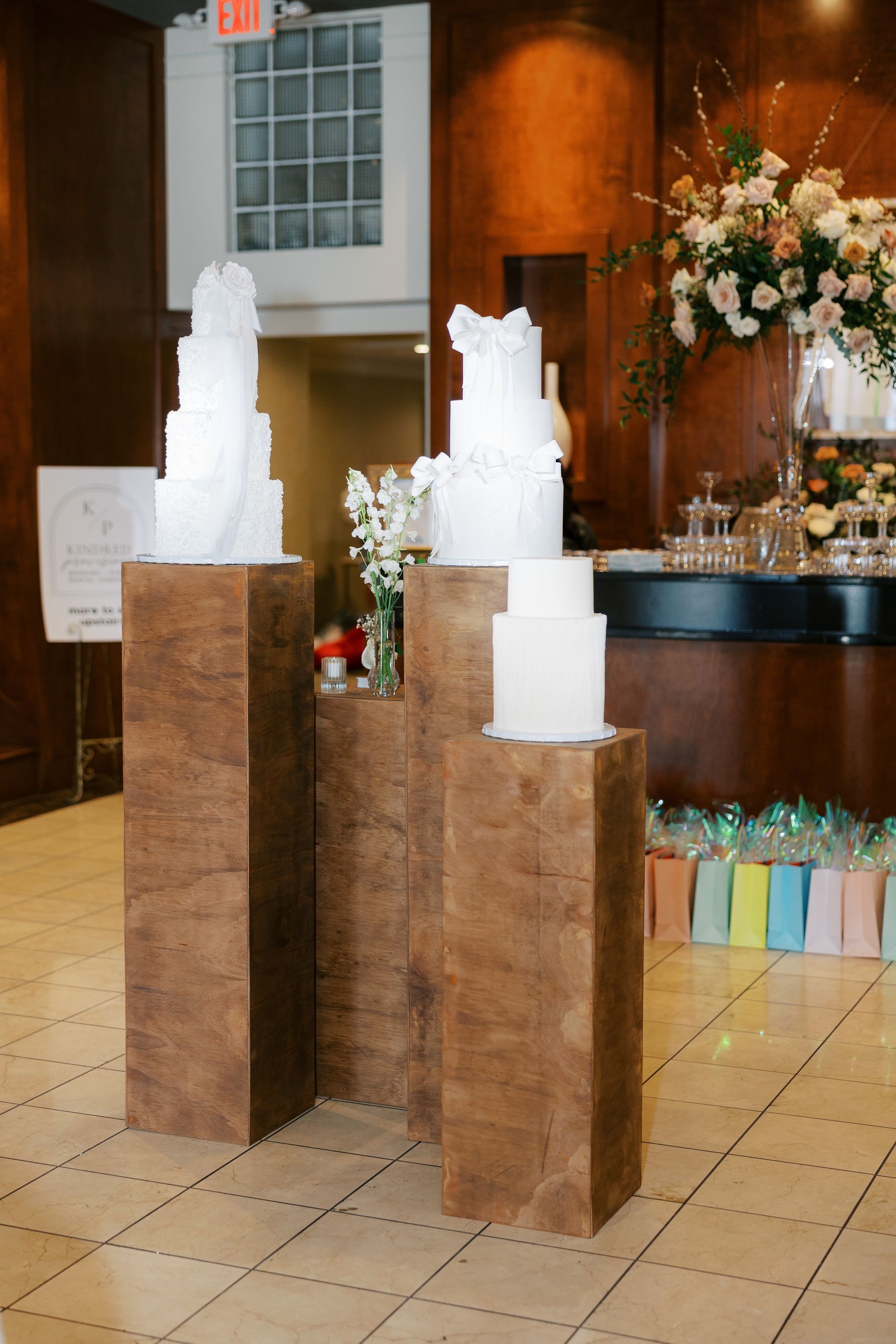 Three white wedding cakes on brown pedestals, indoors. Flowers and decorations nearby.