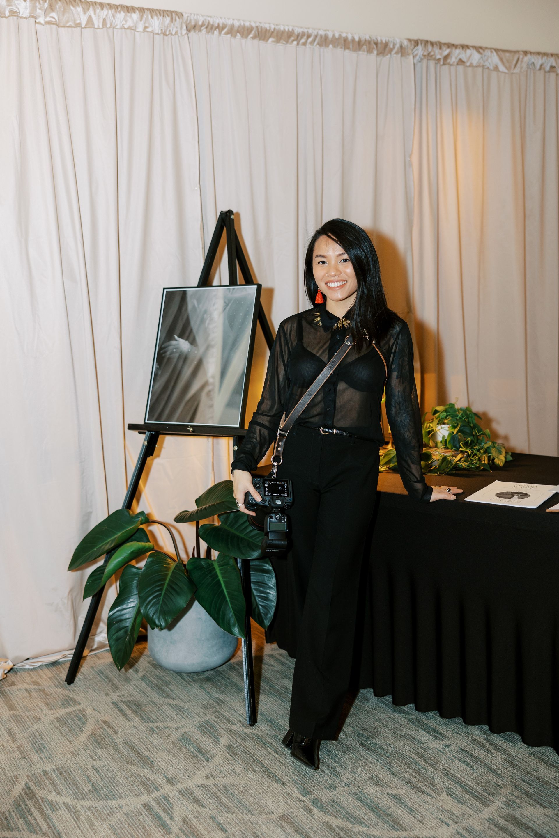 Woman in black outfit stands near a table and painting on easel. She wears red earrings.