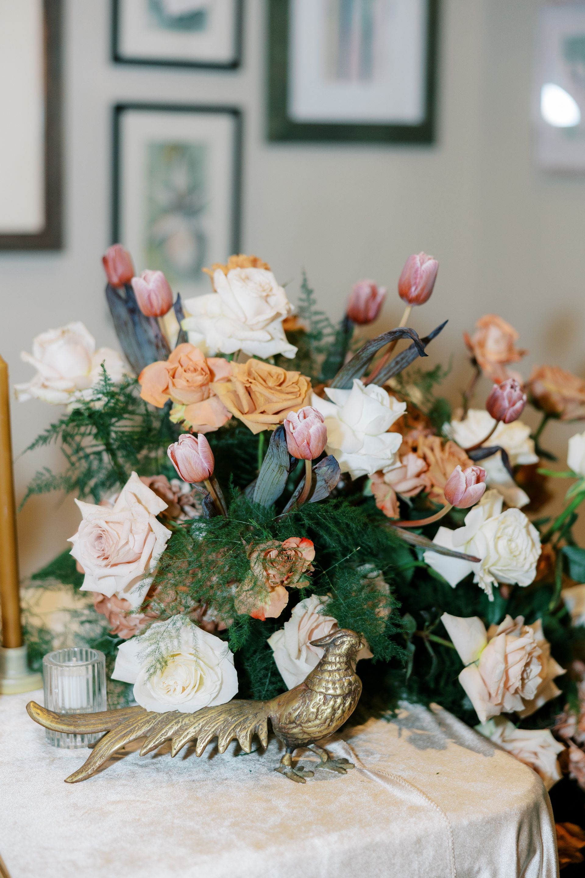 Floral arrangement with tulips, roses, and greenery, on a table with gold pheasant figurine, framed art in background.