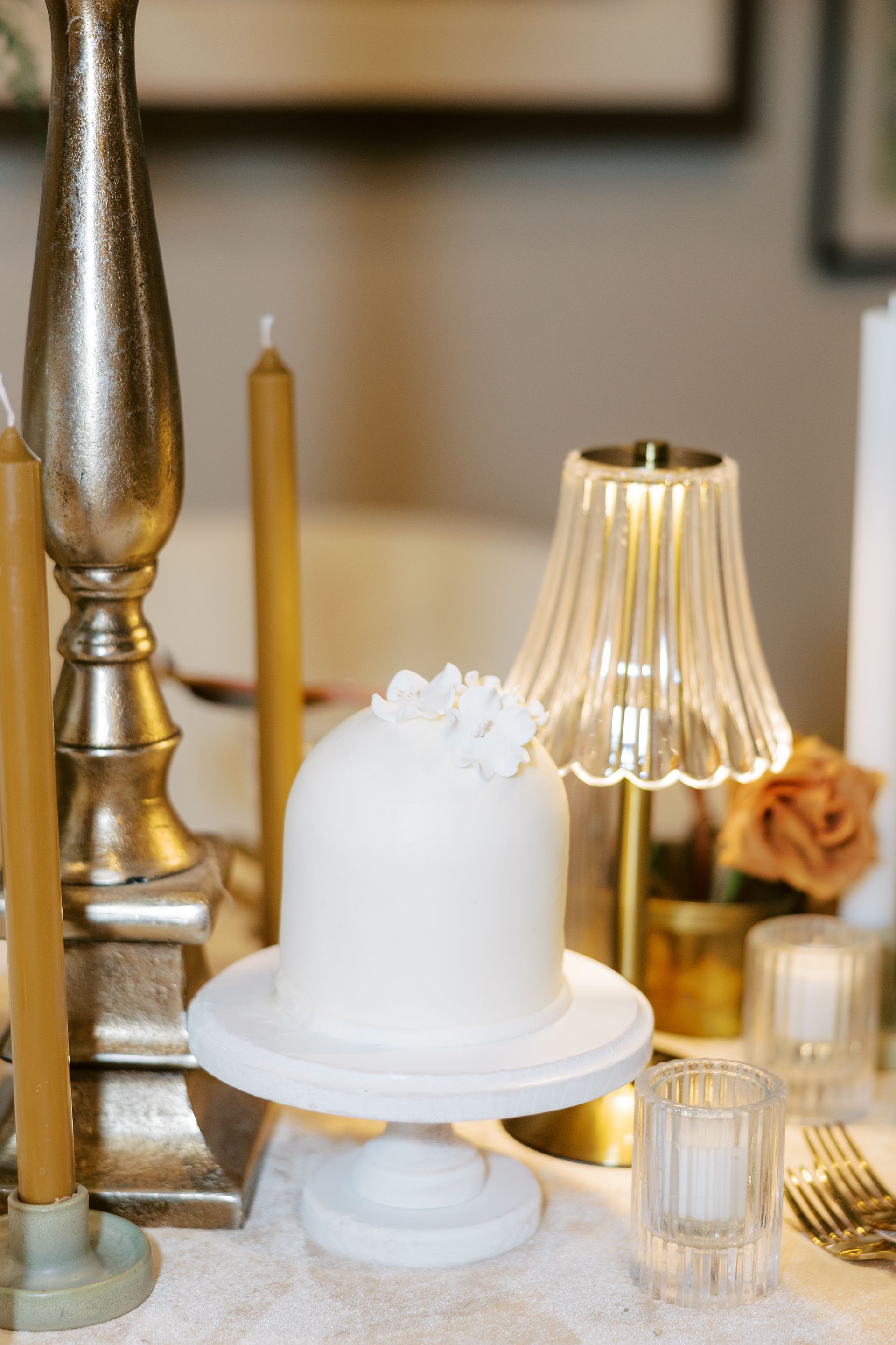 White cake on marble pedestal with candles and a lamp on a table.