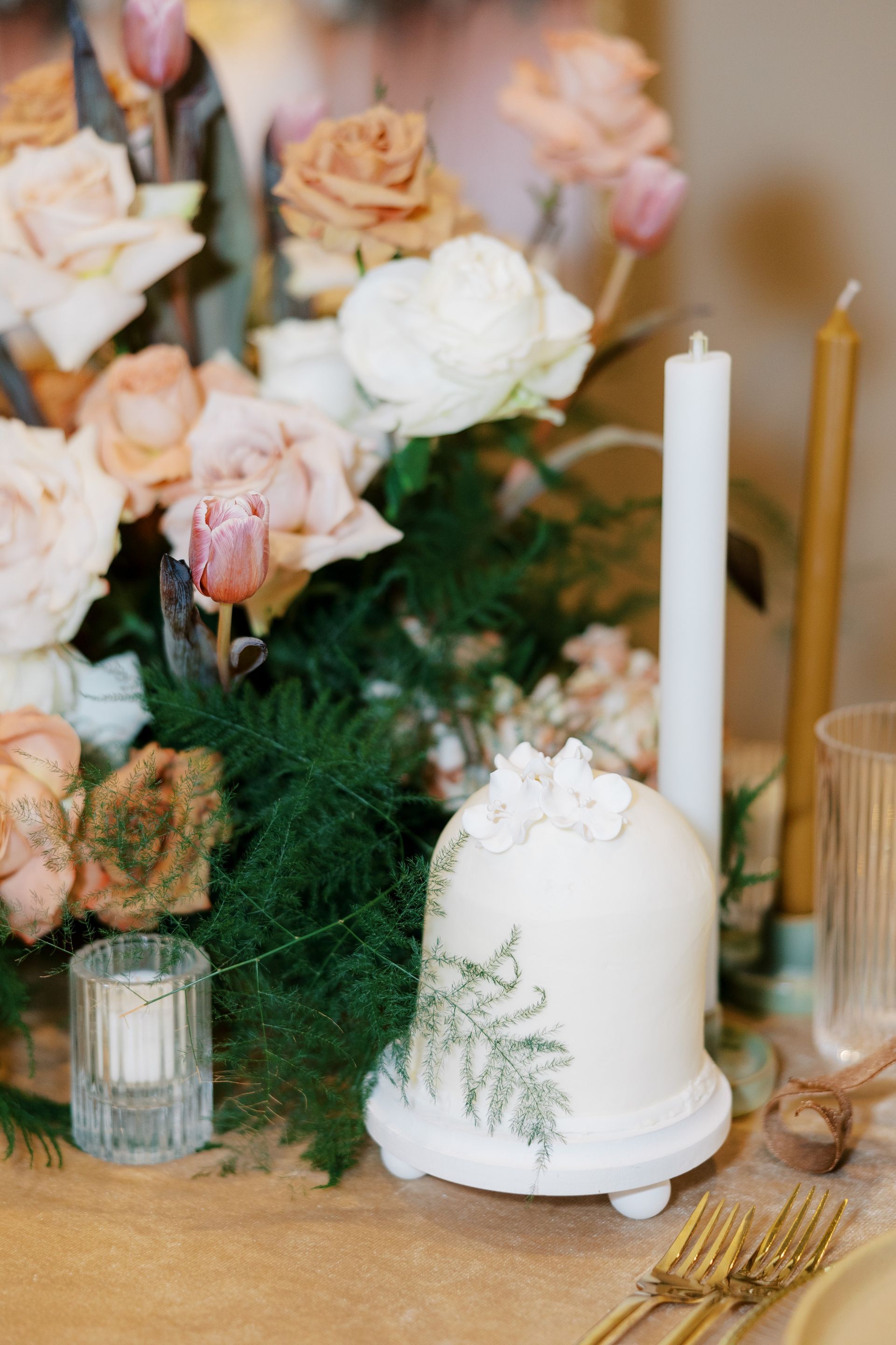 Floral centerpiece with small cake under a cloche, candles, and gold flatware on a table.