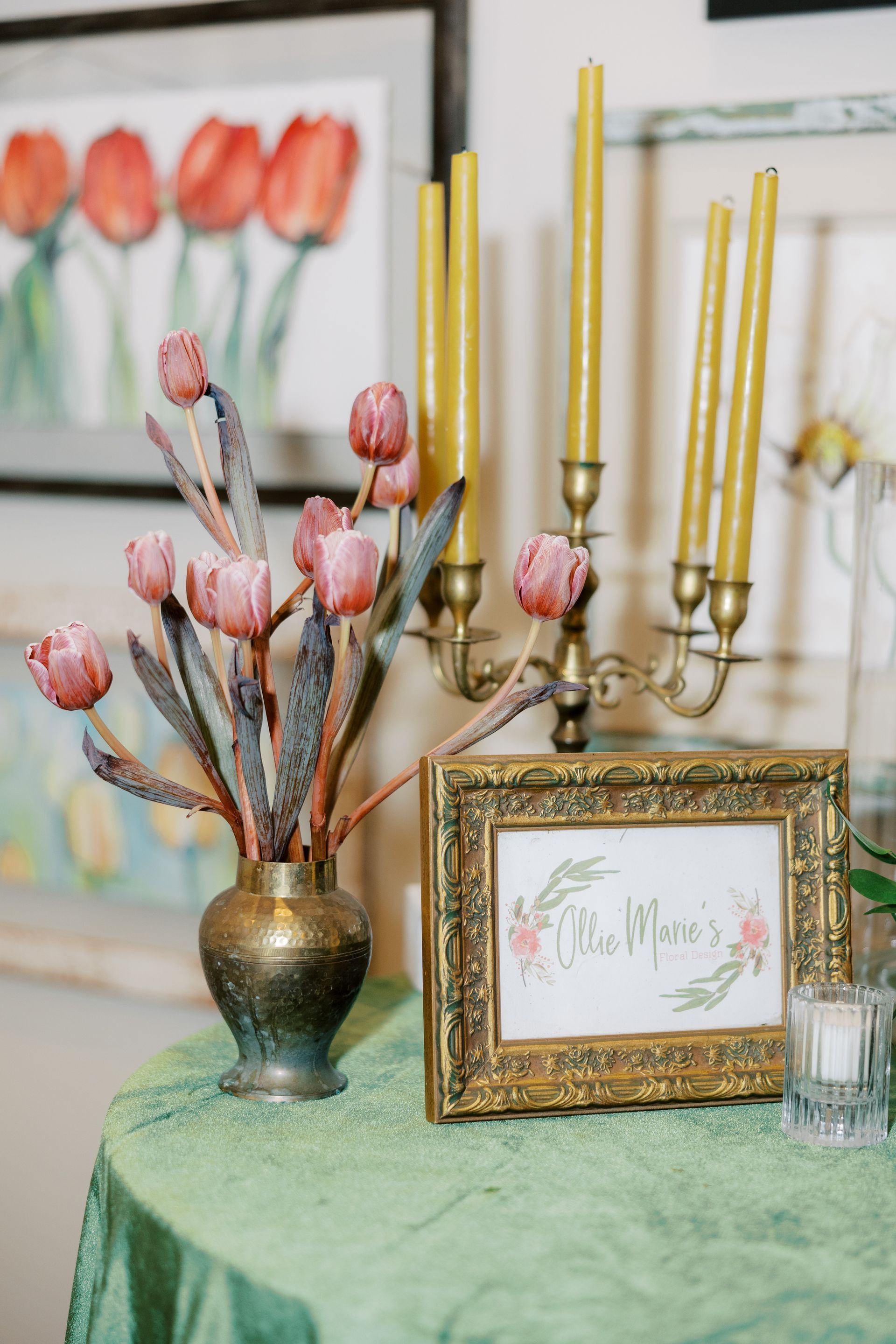 Pink tulips in vase with candelabra and framed floral design on green tablecloth.