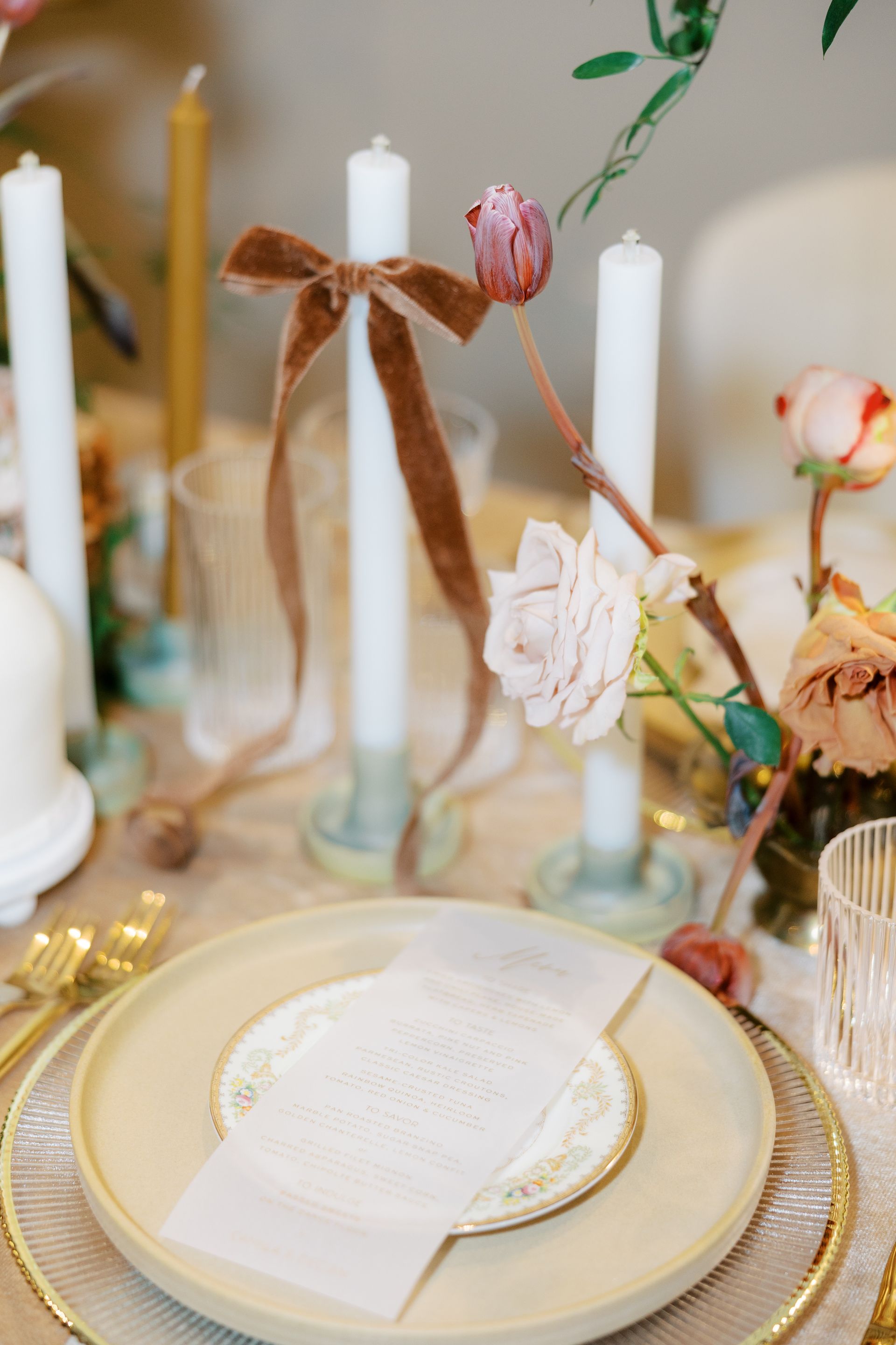 Elegant table setting with candles, flowers, and a menu. Beige, gold, and brown tones.