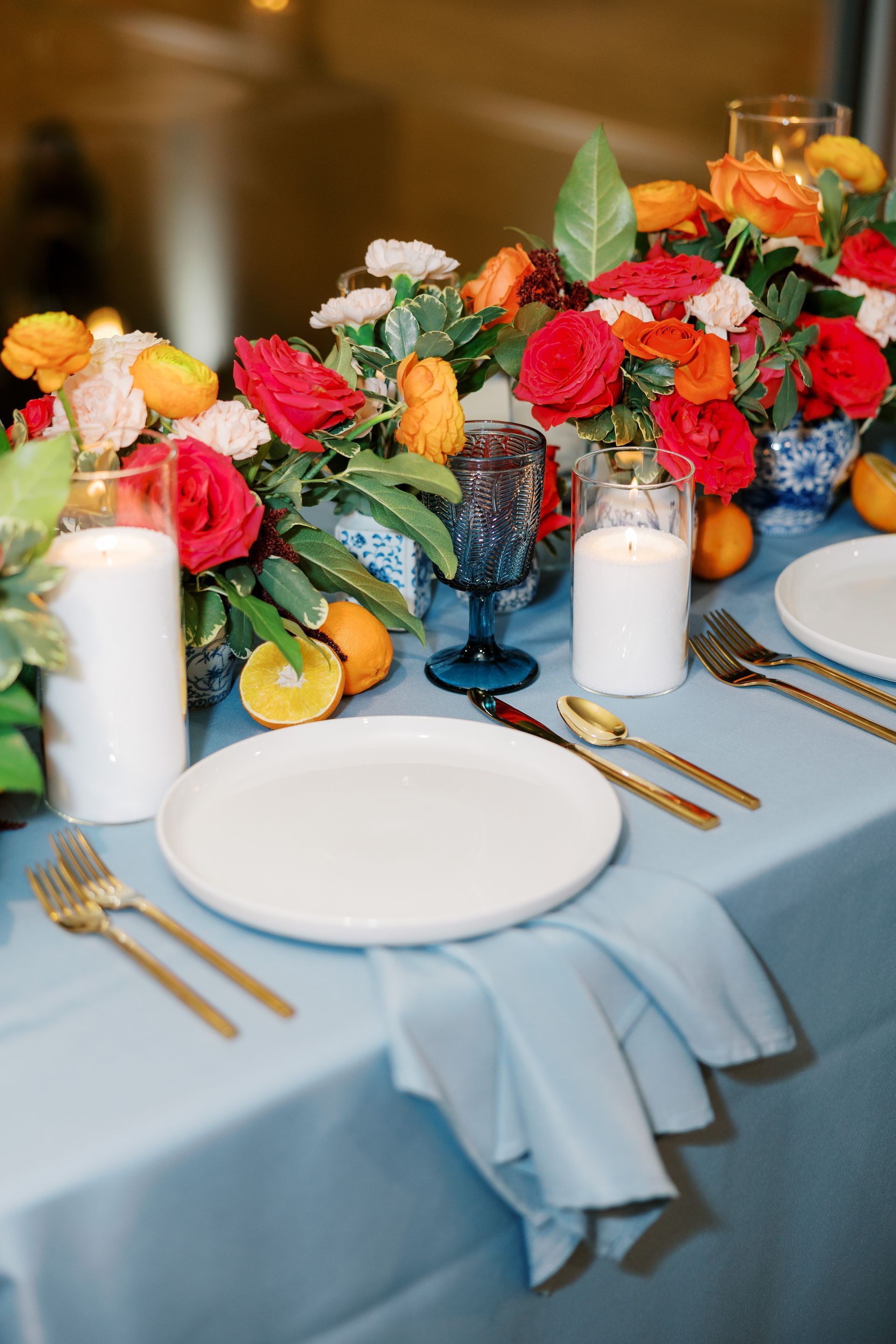 Table setting with blue tablecloth, white plates, gold flatware, floral centerpiece, and candles.