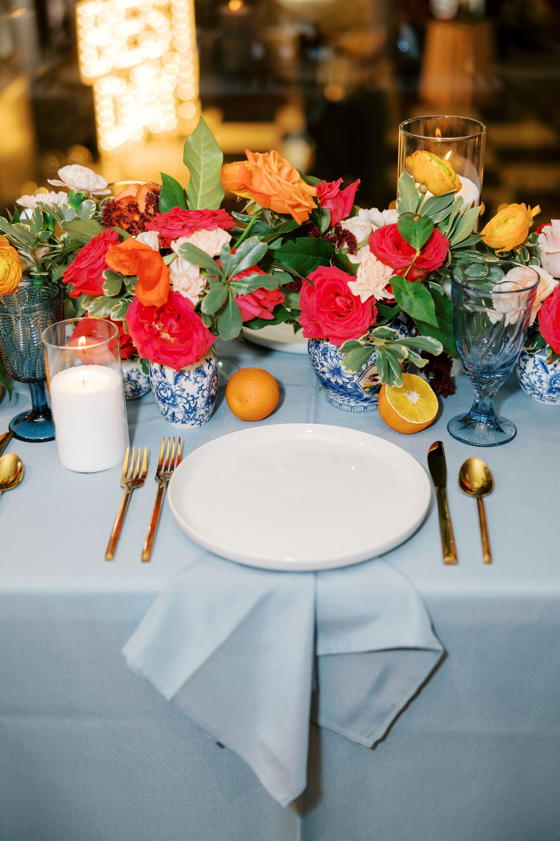 Elegant table setting with colorful floral arrangement, gold cutlery, blue glasses, and a white plate on a light blue tablecloth.