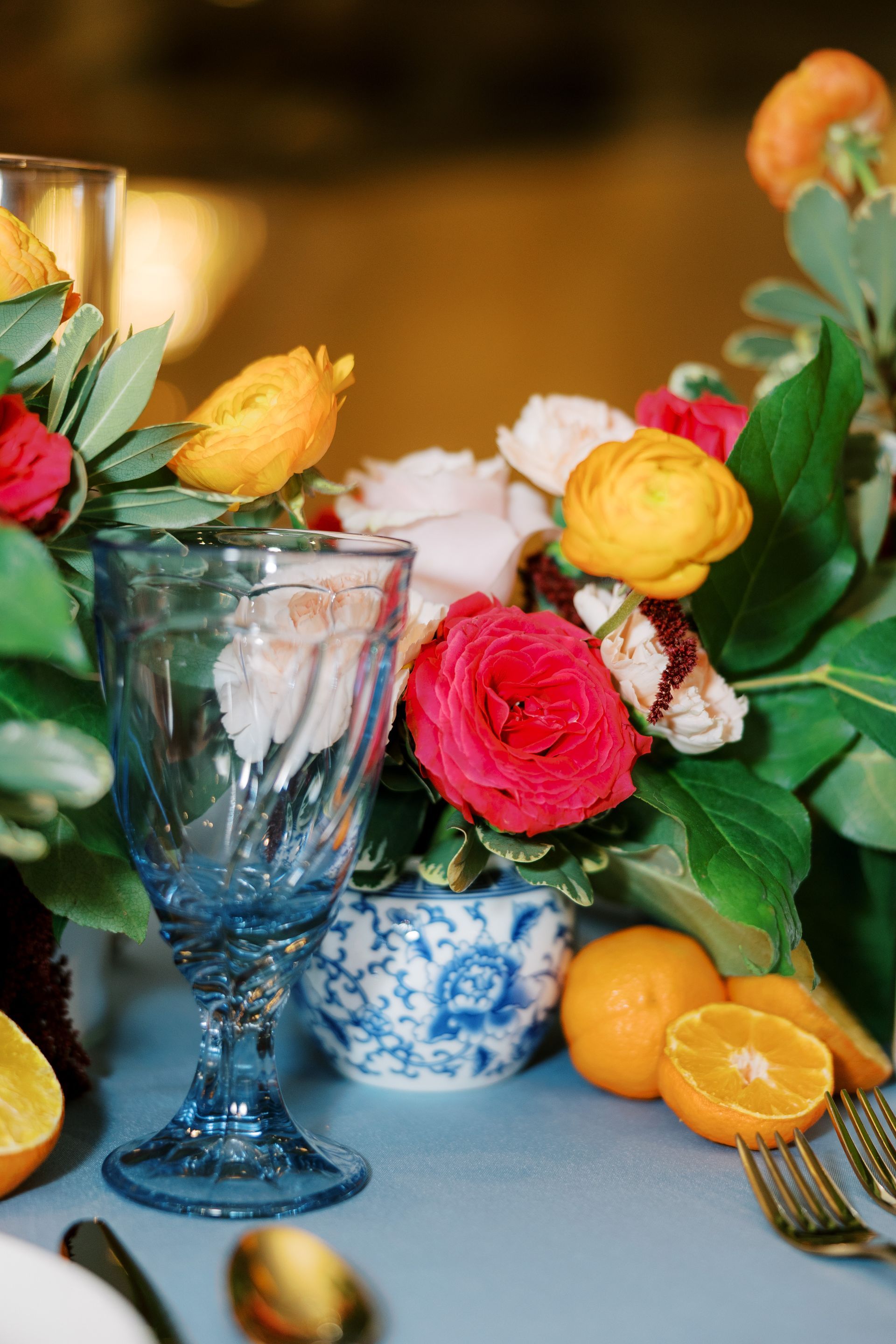 Blue-tinted glass, floral centerpiece with roses, oranges, and gold silverware on a blue tablecloth.