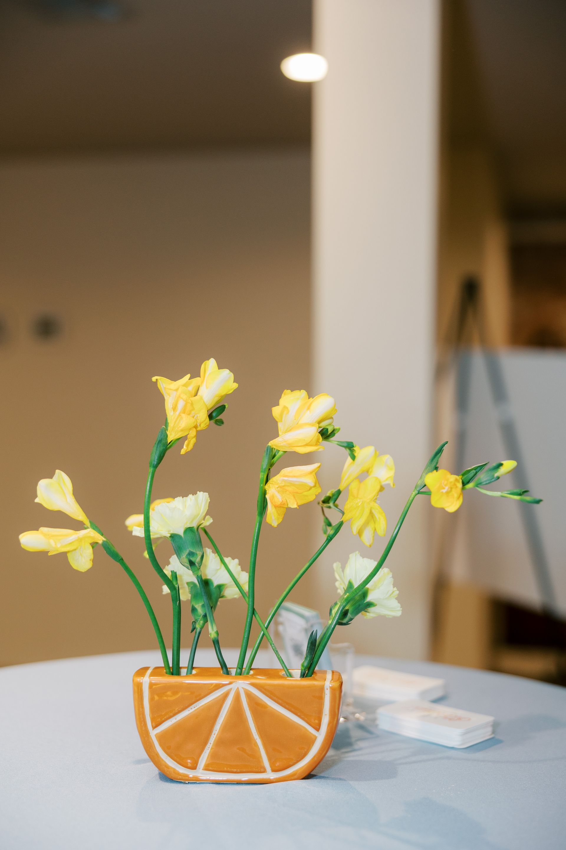 Yellow flowers in a gingerbread orange slice-shaped vase on a light blue tablecloth.