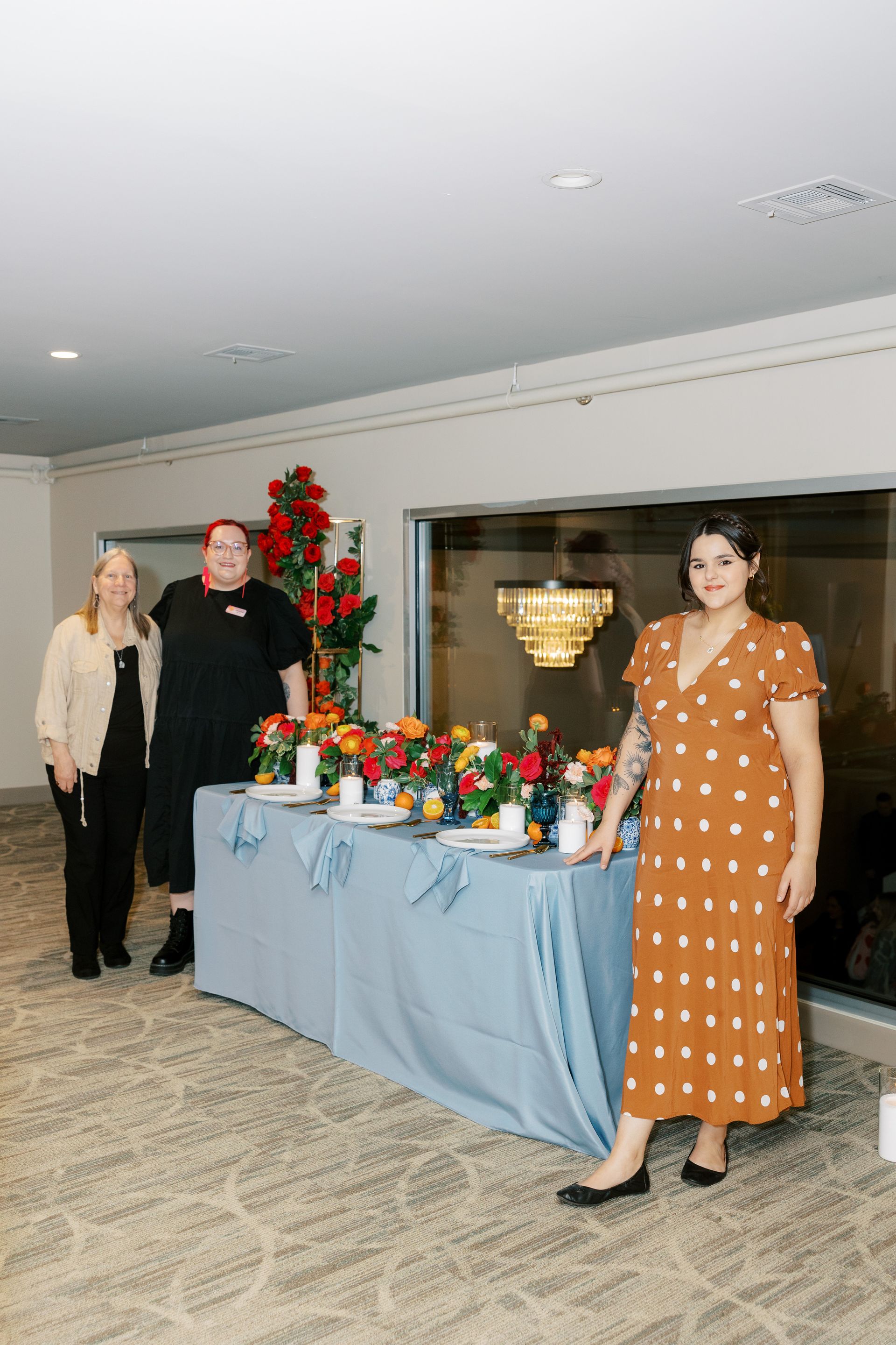 Three people stand near a table with flowers. One in a brown dress, the others in black. Window behind.
