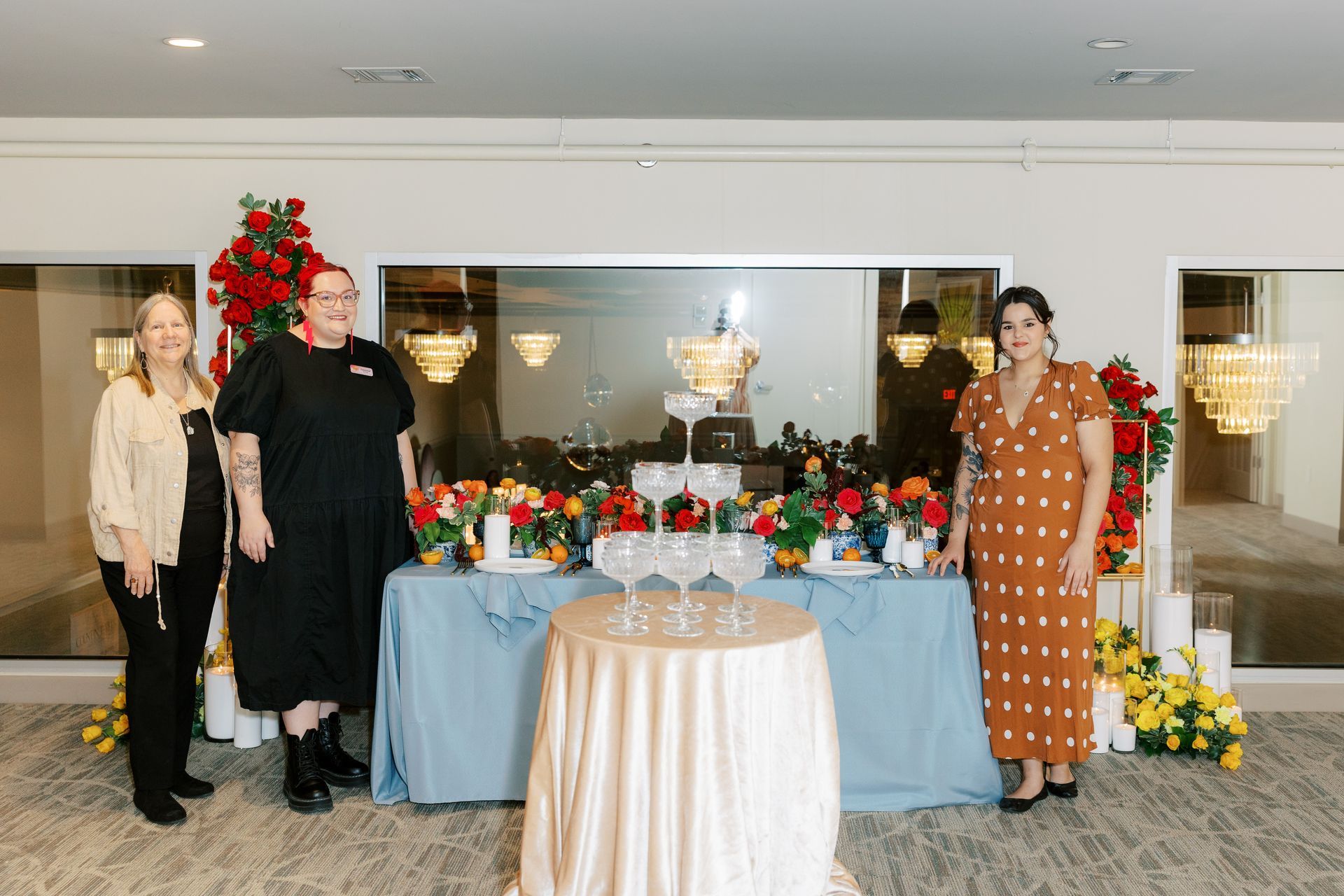 Three women pose near a table with flowers, champagne glasses, and a cake. A large window and chandeliers are in the background.