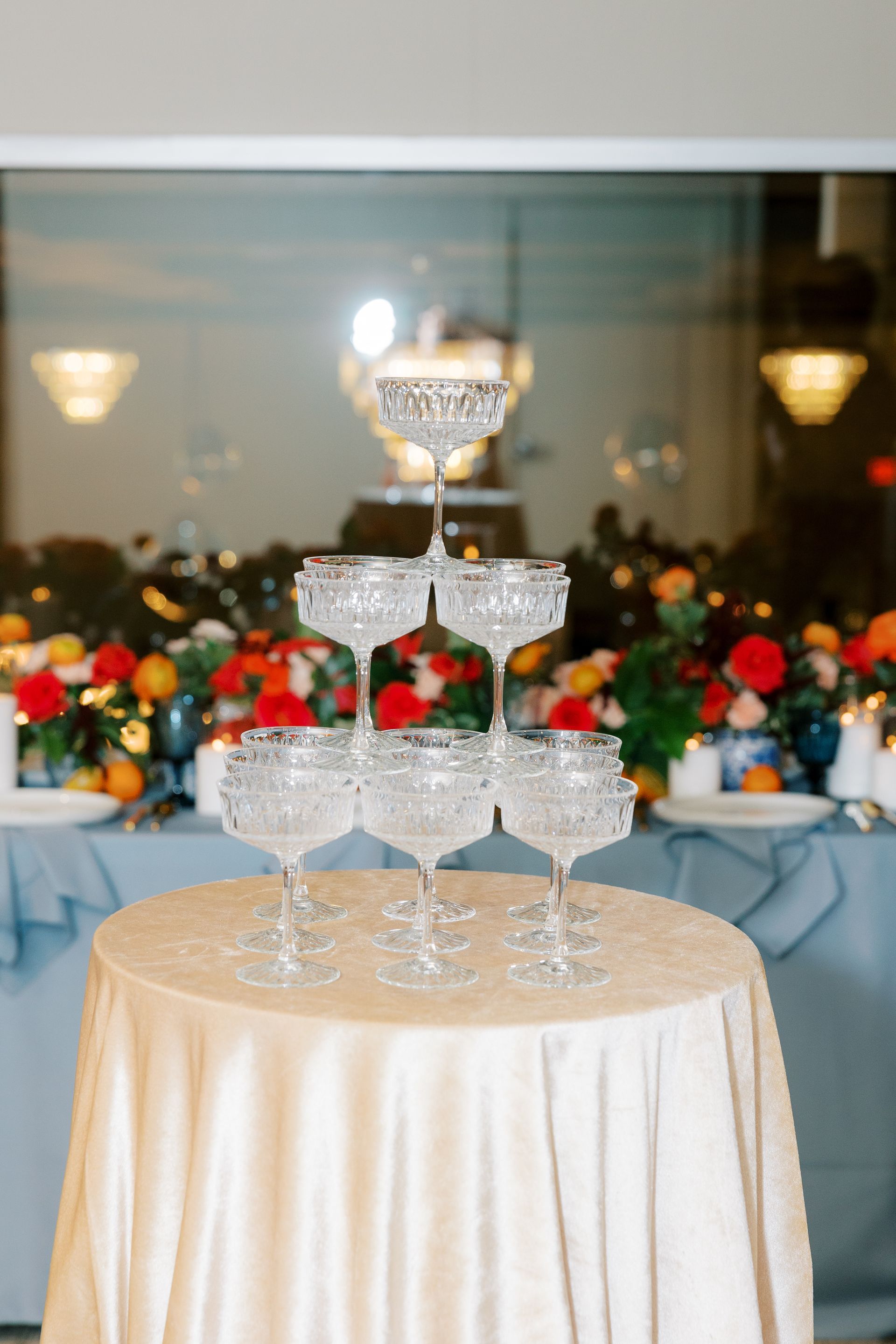 Champagne glasses stacked in a pyramid on a table at an event, with floral decorations in the background.