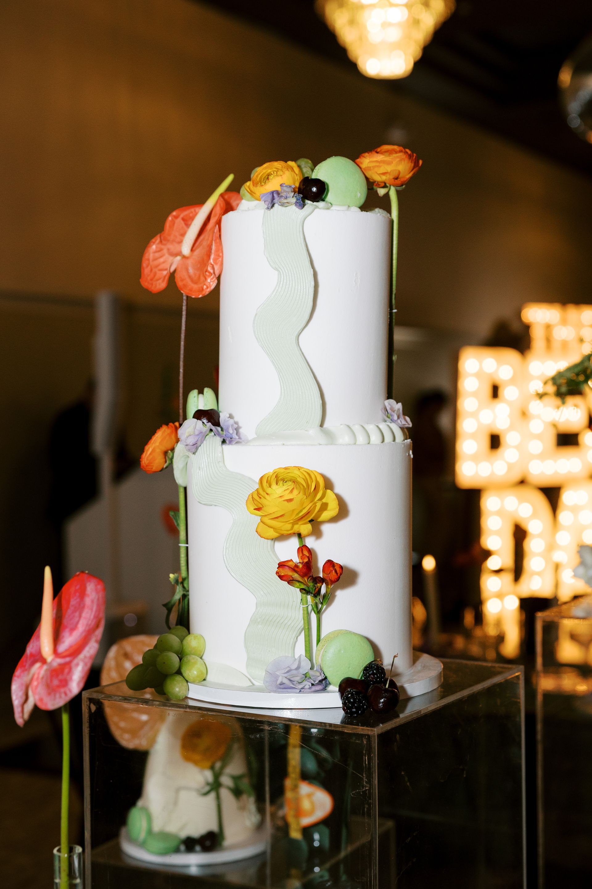 Two-tiered white cake decorated with flowers, macaroons, and a light green wavy design, displayed on a clear stand.