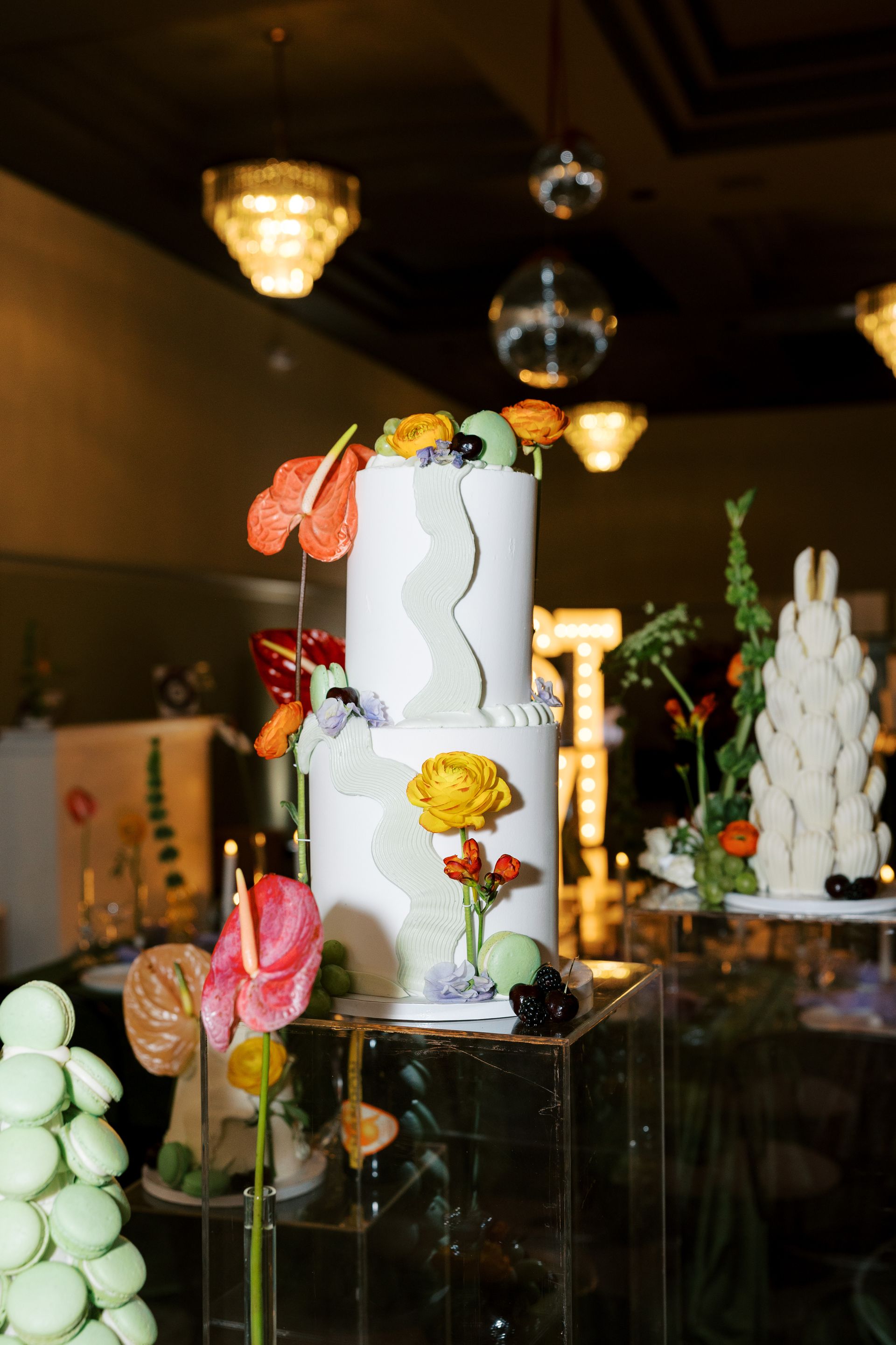 Two-tiered white cake with colorful floral decor on a clear stand, surrounded by more desserts and flowers in a room.