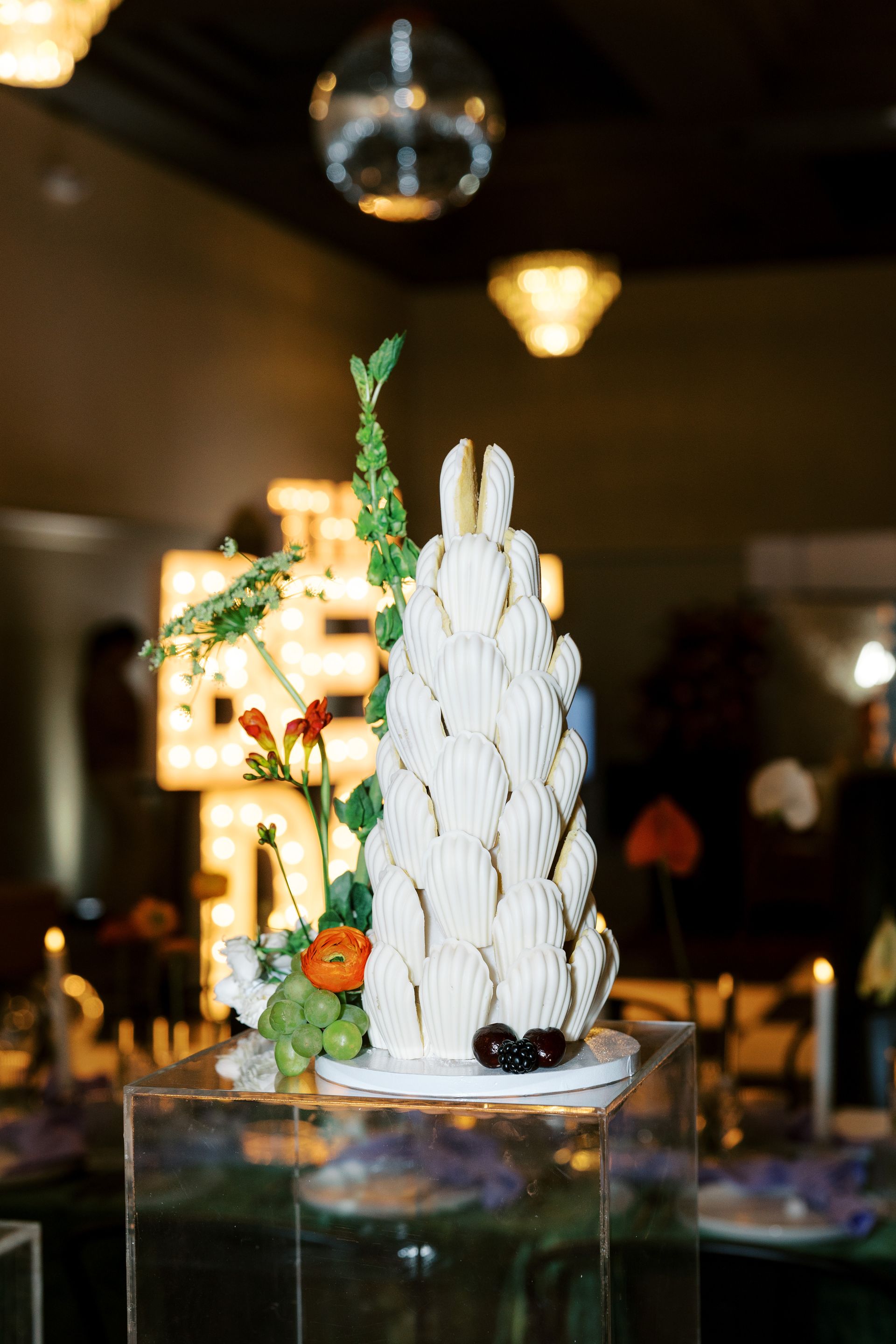 Towering white dessert decorated with flowers, atop a clear pedestal, with a disco ball and other decor in the background.