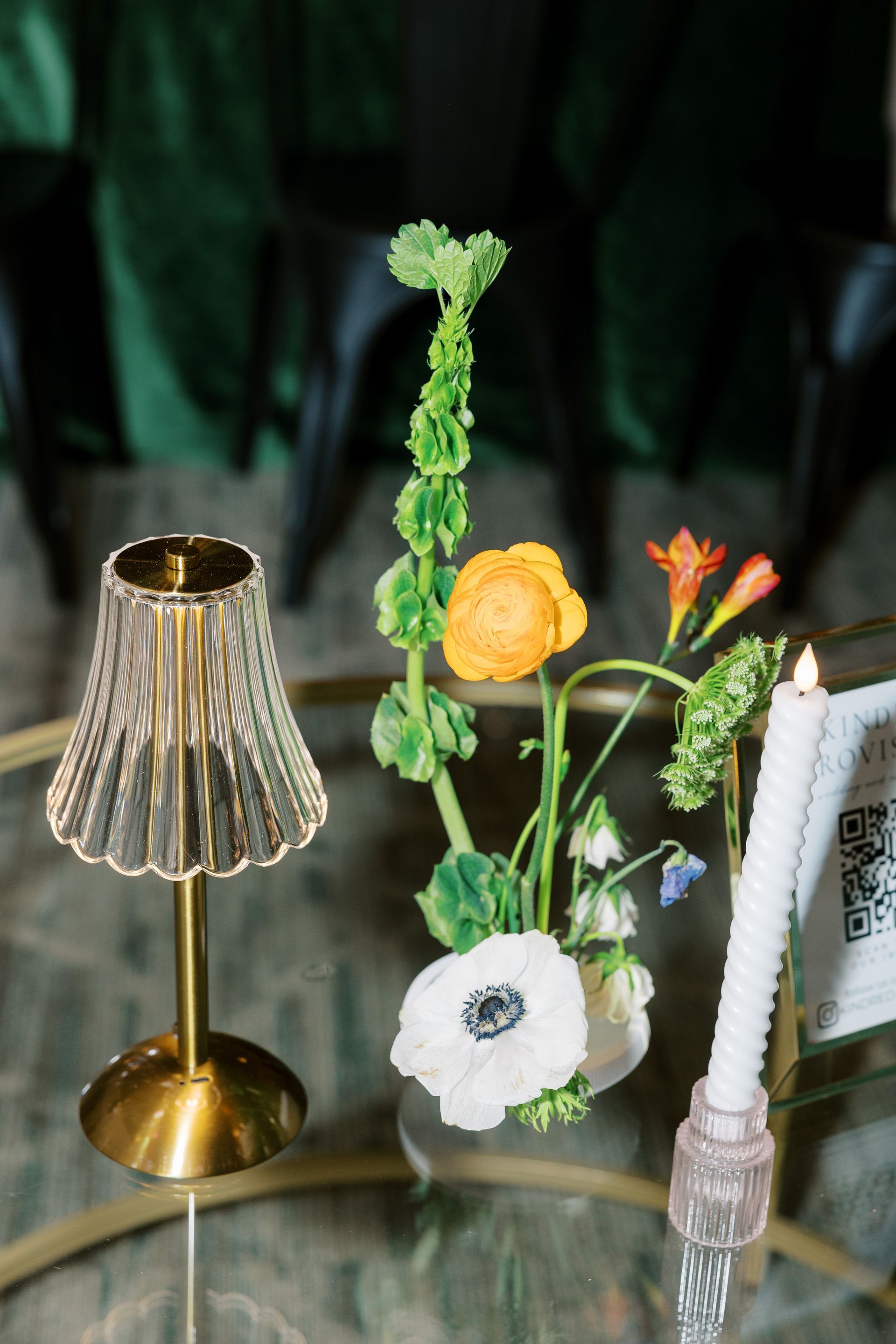 A gold lamp and flower arrangement on a glass table. A lit candle sits nearby.